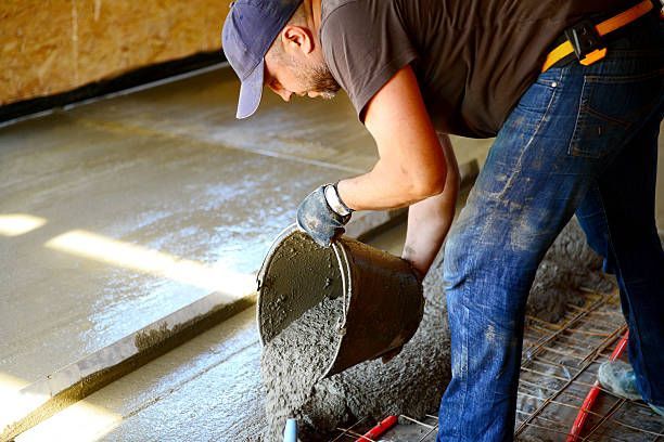 Person pours concrete from a bucket onto a floor under construction; brown, blue, and grey colors.