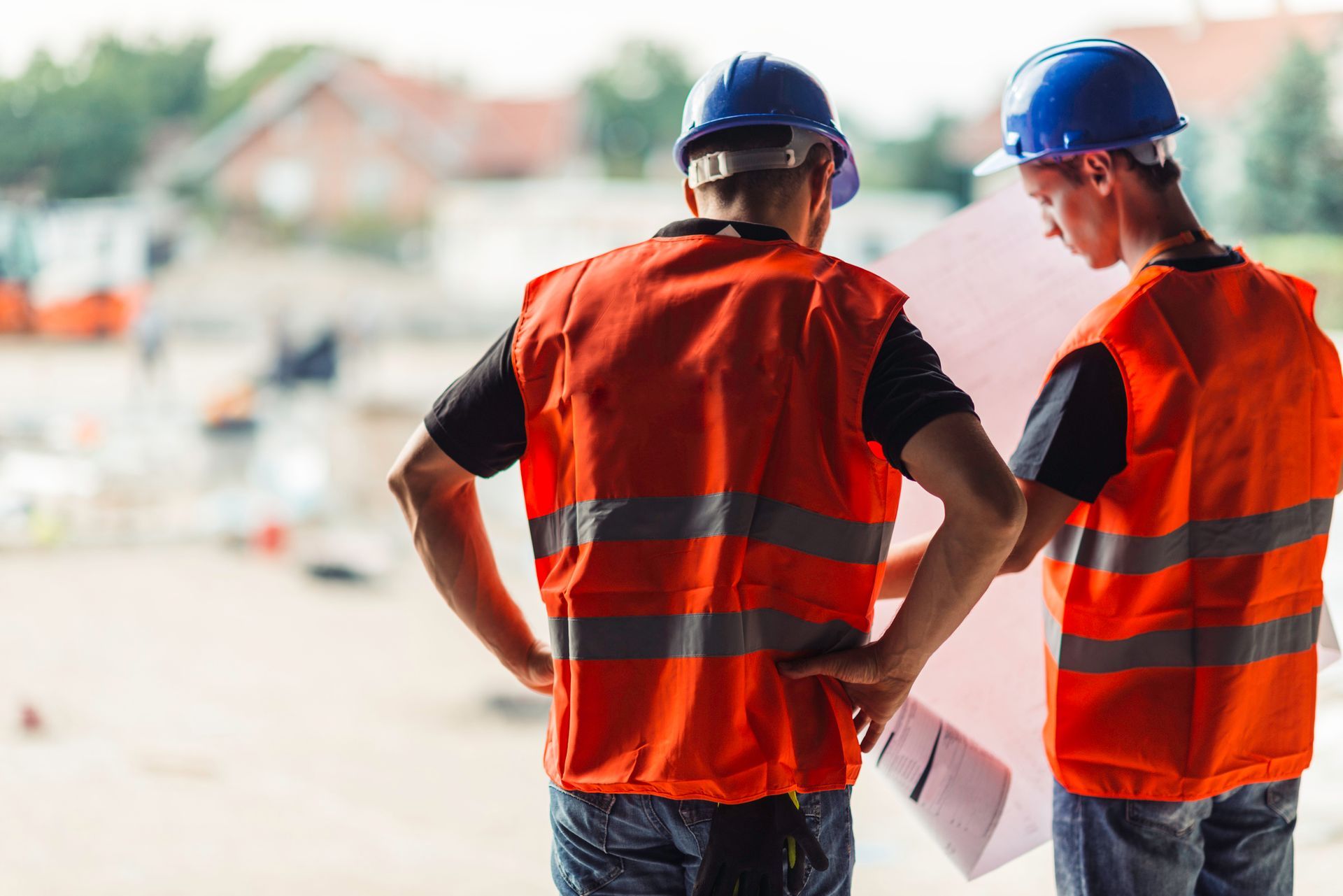Two construction workers in vests and hard hats reviewing blueprints outdoors.