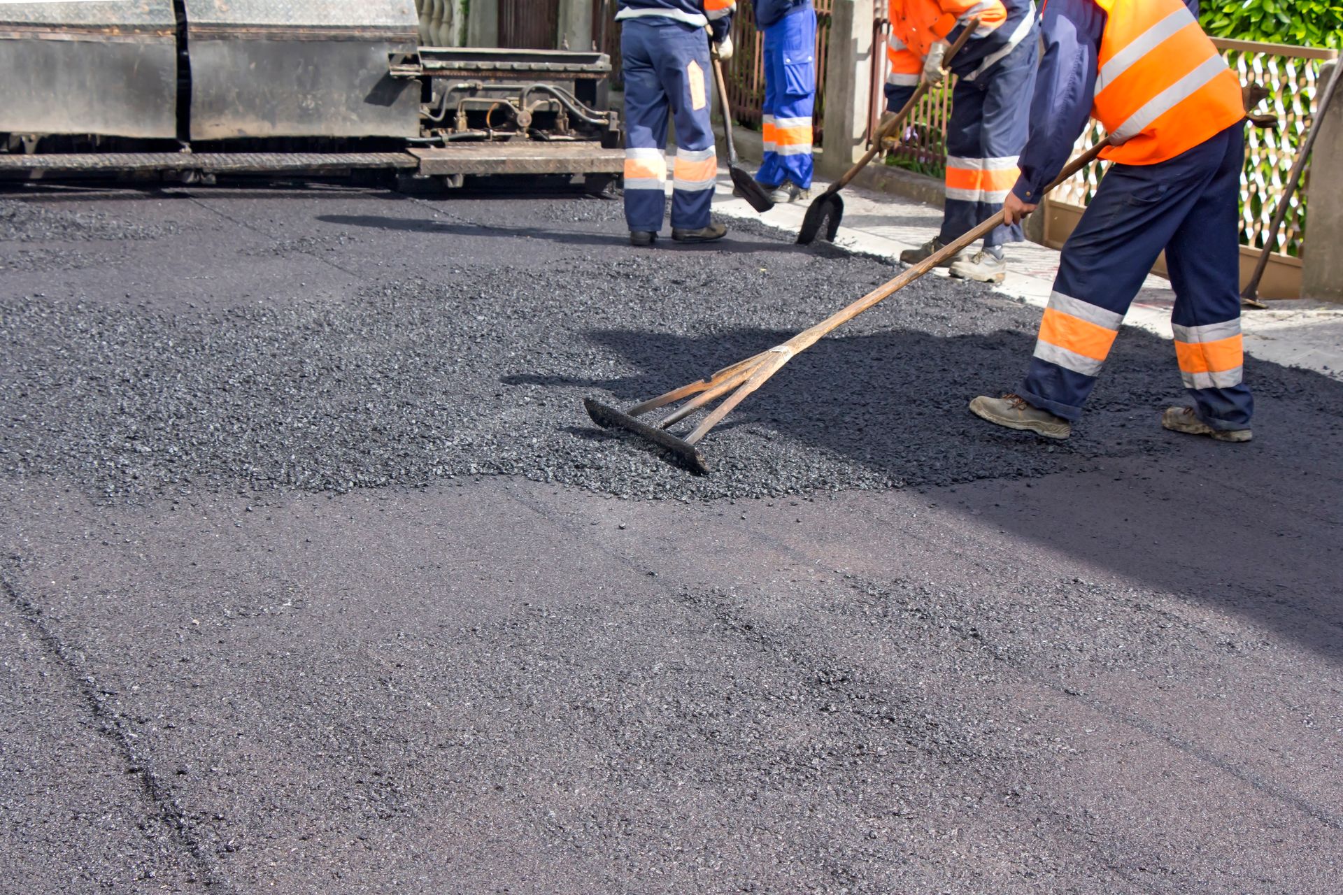 Construction workers paving a road, using rakes on fresh asphalt, wearing reflective vests.