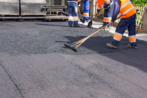 Construction workers paving a road, using rakes on fresh asphalt, wearing reflective vests.