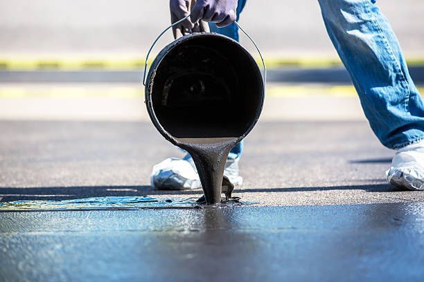 Person pouring black sealant from a bucket onto a paved surface.