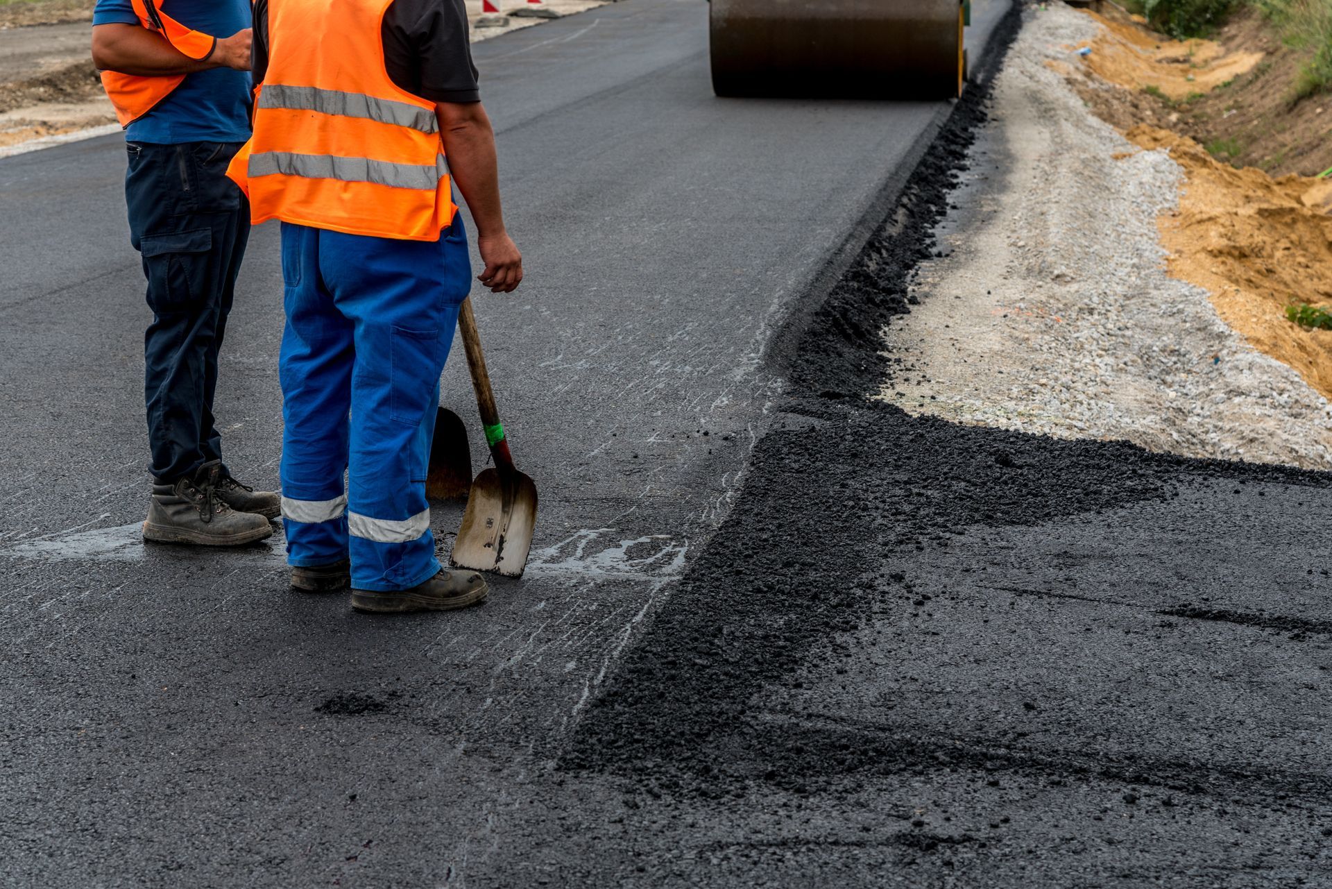 Two road workers in safety vests stand near freshly laid asphalt.