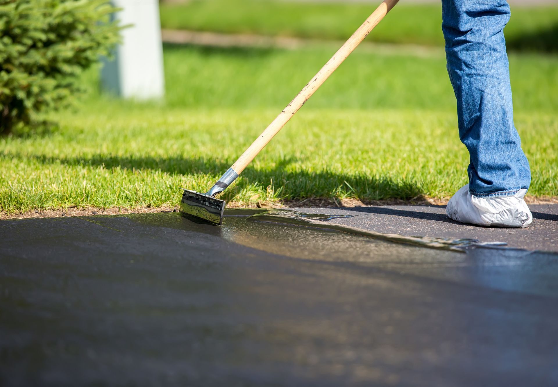 Person sealing a driveway with a long-handled brush; black asphalt, green grass.