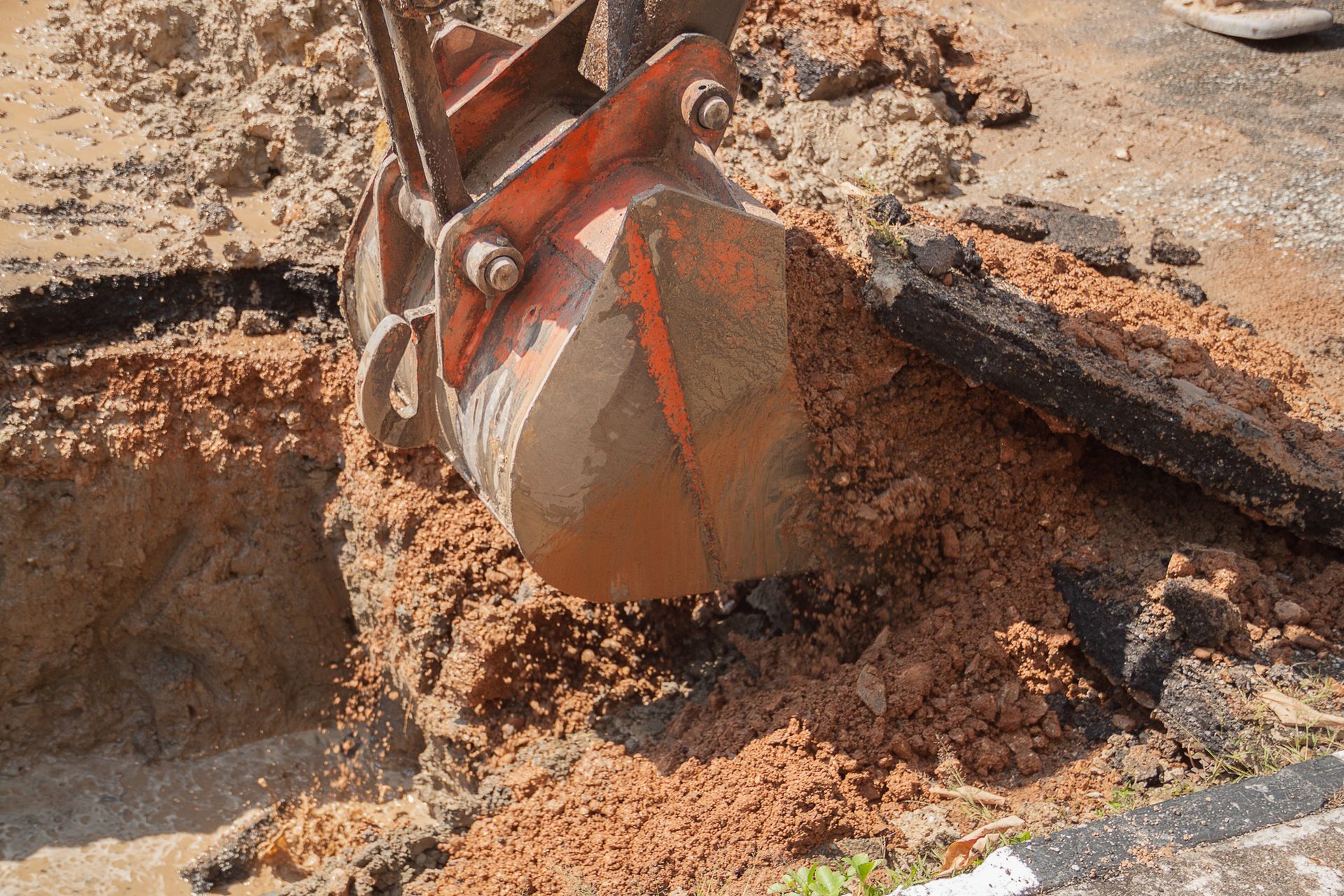 An excavator bucket digging into dirt and asphalt, showing a construction site.