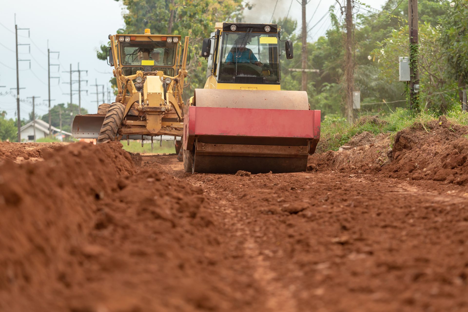 Road construction: a grader and roller compacting red dirt. Power lines and trees in the background.