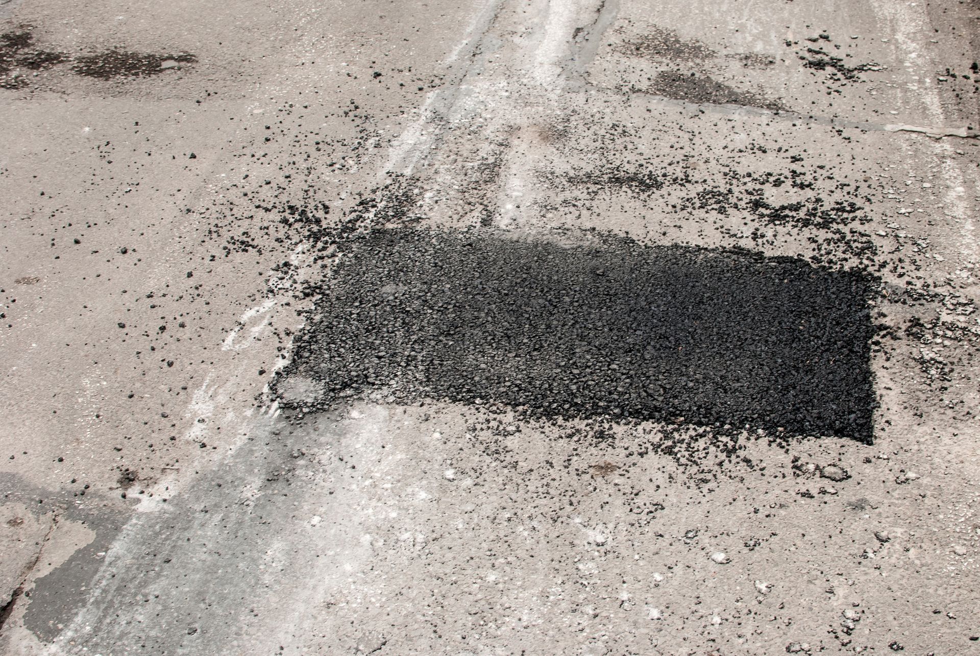 Asphalt patch on a concrete road surface, with loose asphalt debris scattered around.