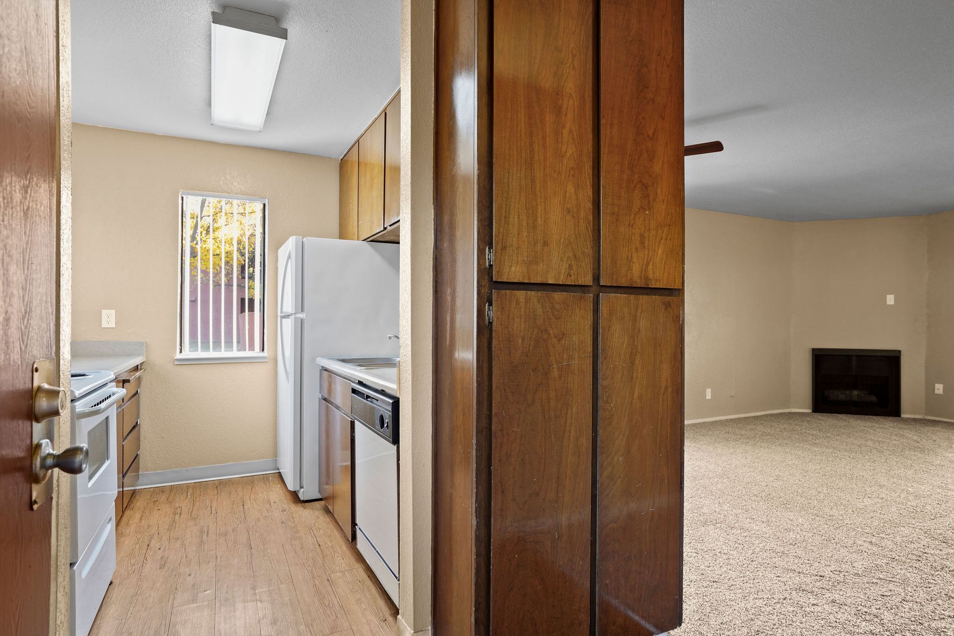 Small kitchen with white appliances and wood cabinets opening to a beige carpeted living room with a fireplace.