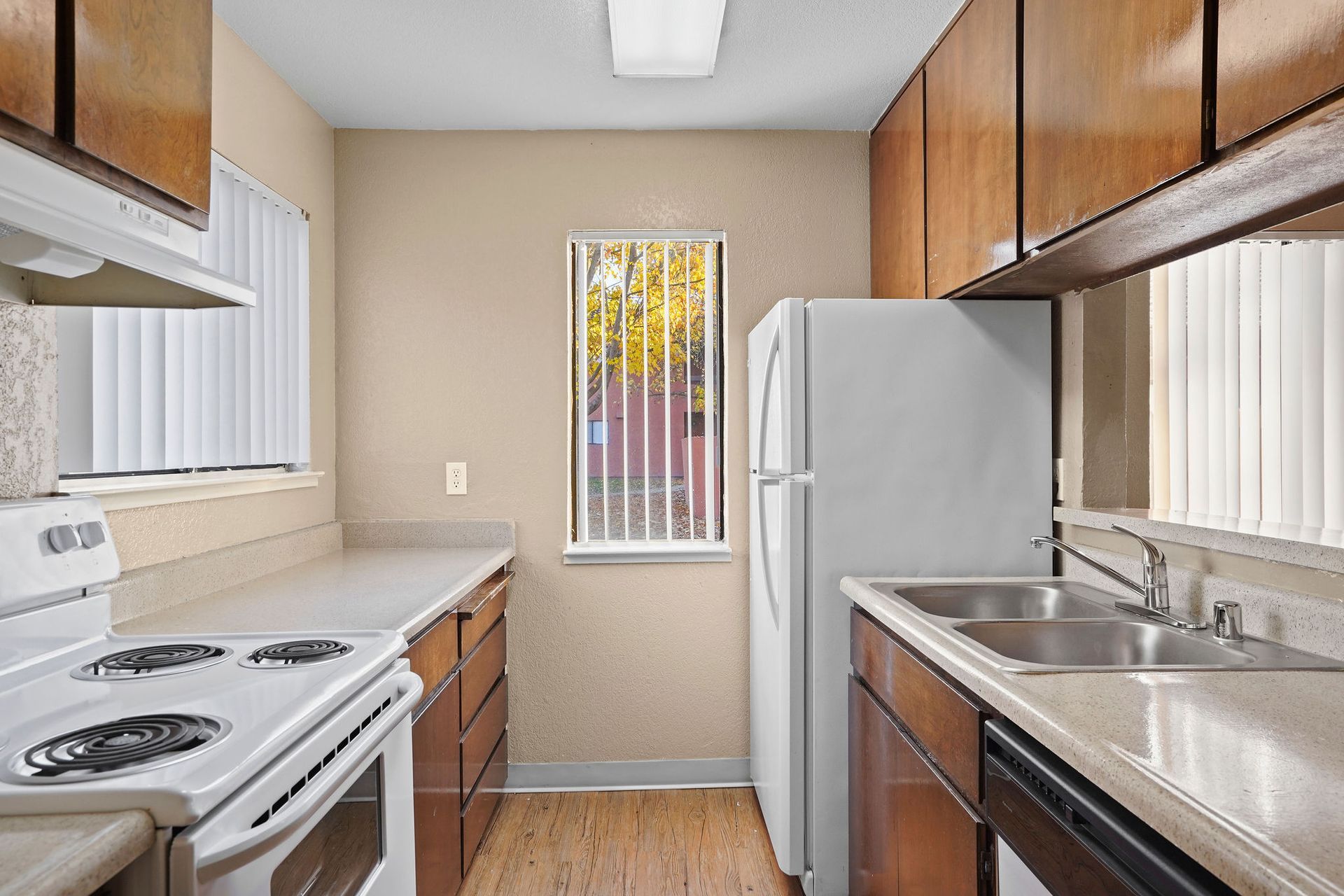 Narrow galley kitchen with white appliances, wooden cabinets, and a window at the end.