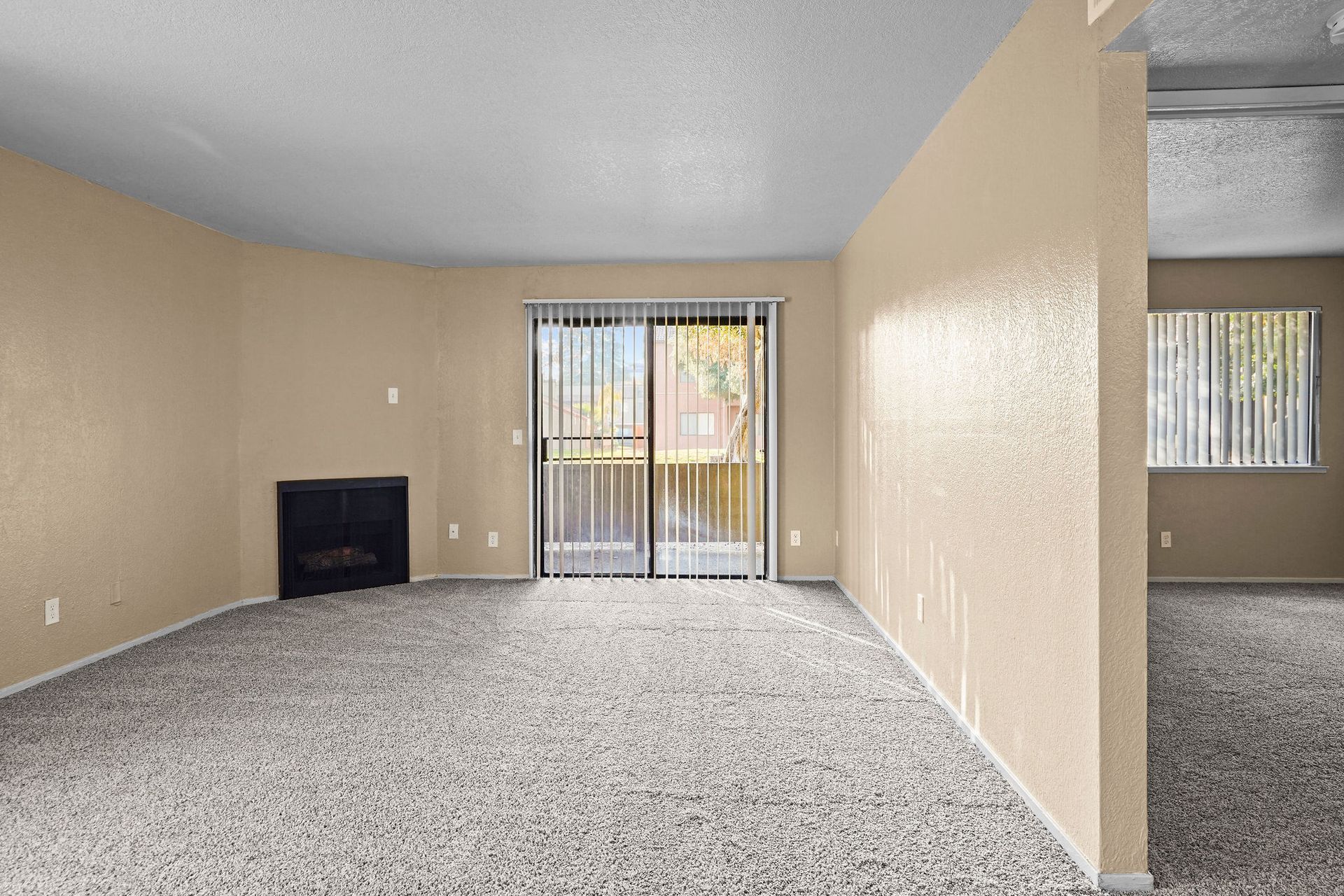 Empty beige living room with gray carpet, fireplace, and sliding glass door to balcony