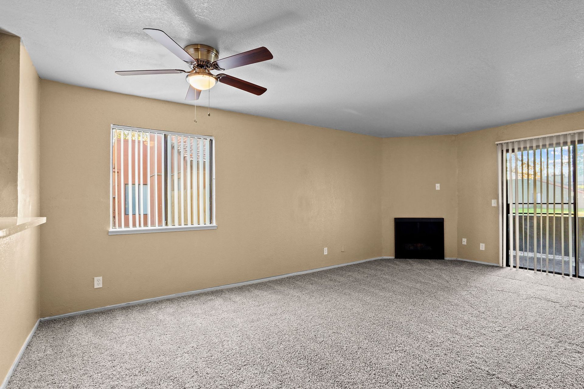 Empty beige living room with gray carpet, ceiling fan, fireplace, and vertical blinds on windows and sliding door
