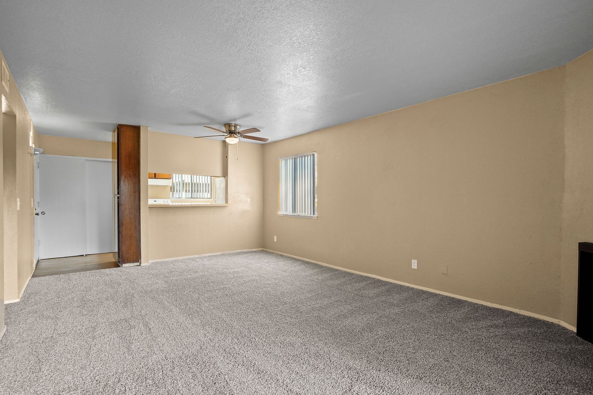 Empty beige living room with gray carpet, ceiling fan, and small kitchen pass-through window