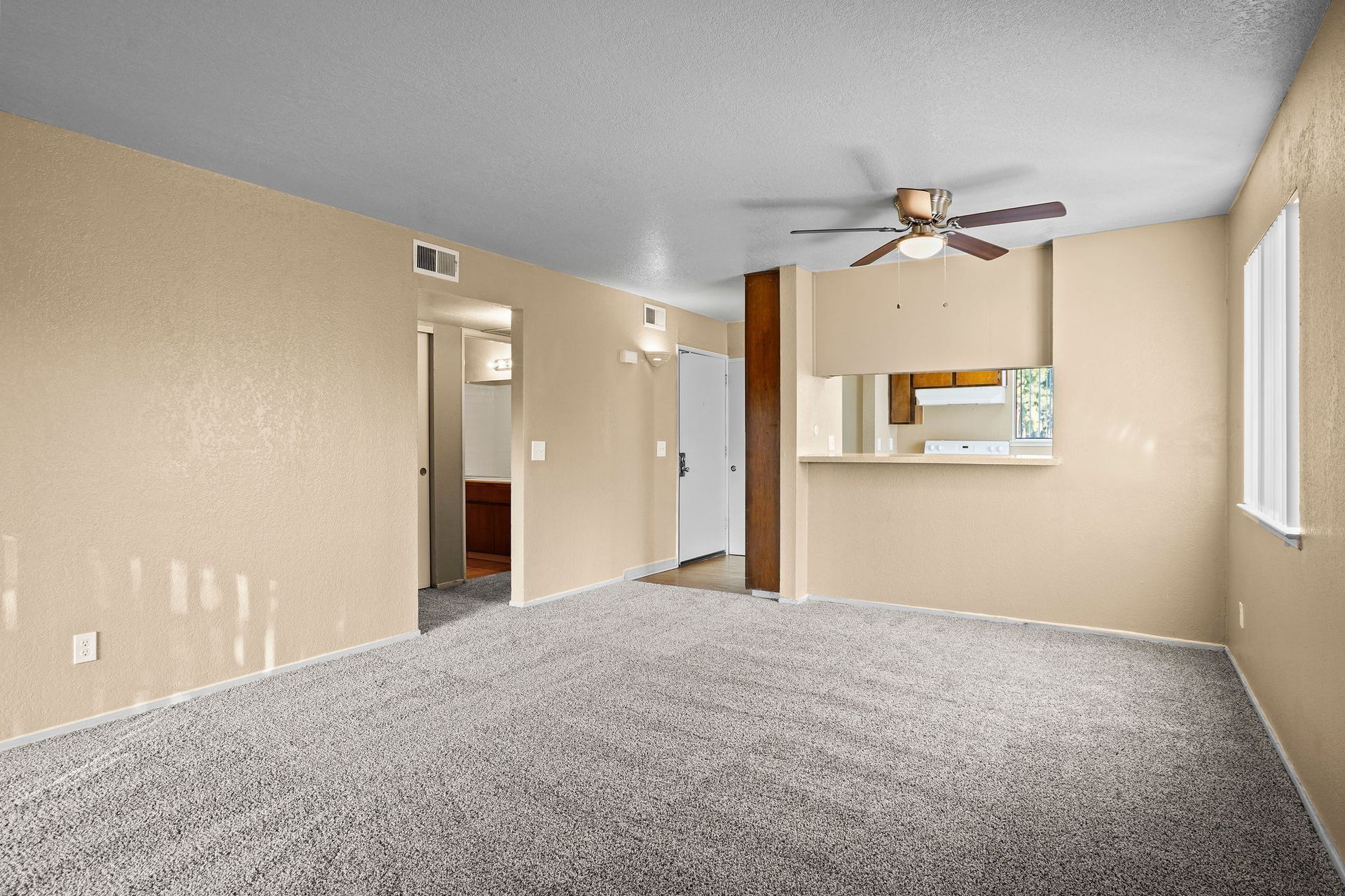 Empty beige living room with gray carpet, ceiling fan, and pass-through window to kitchen