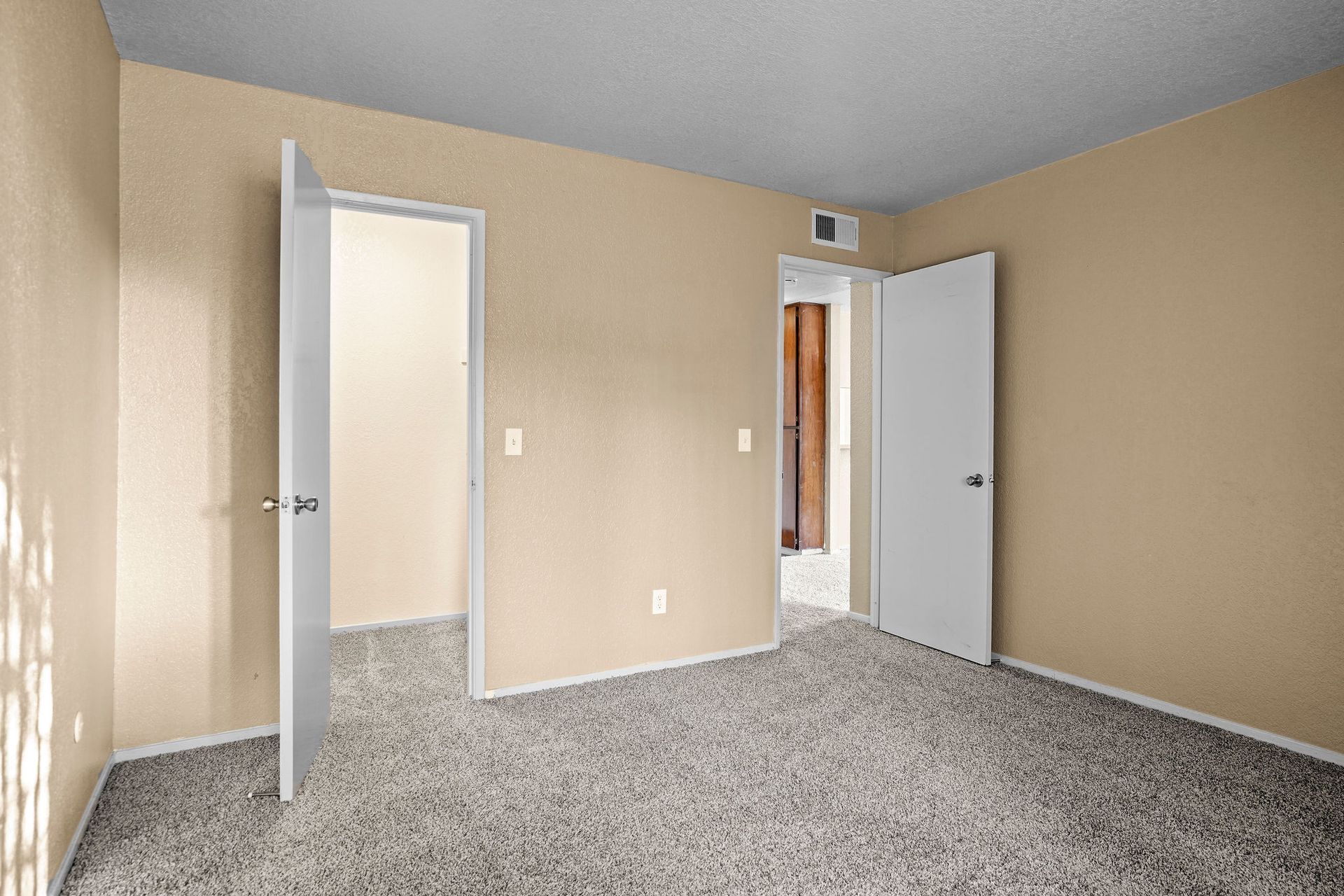 Empty beige bedroom with carpet, two open white doors, and a small wall-mounted device above the doorway