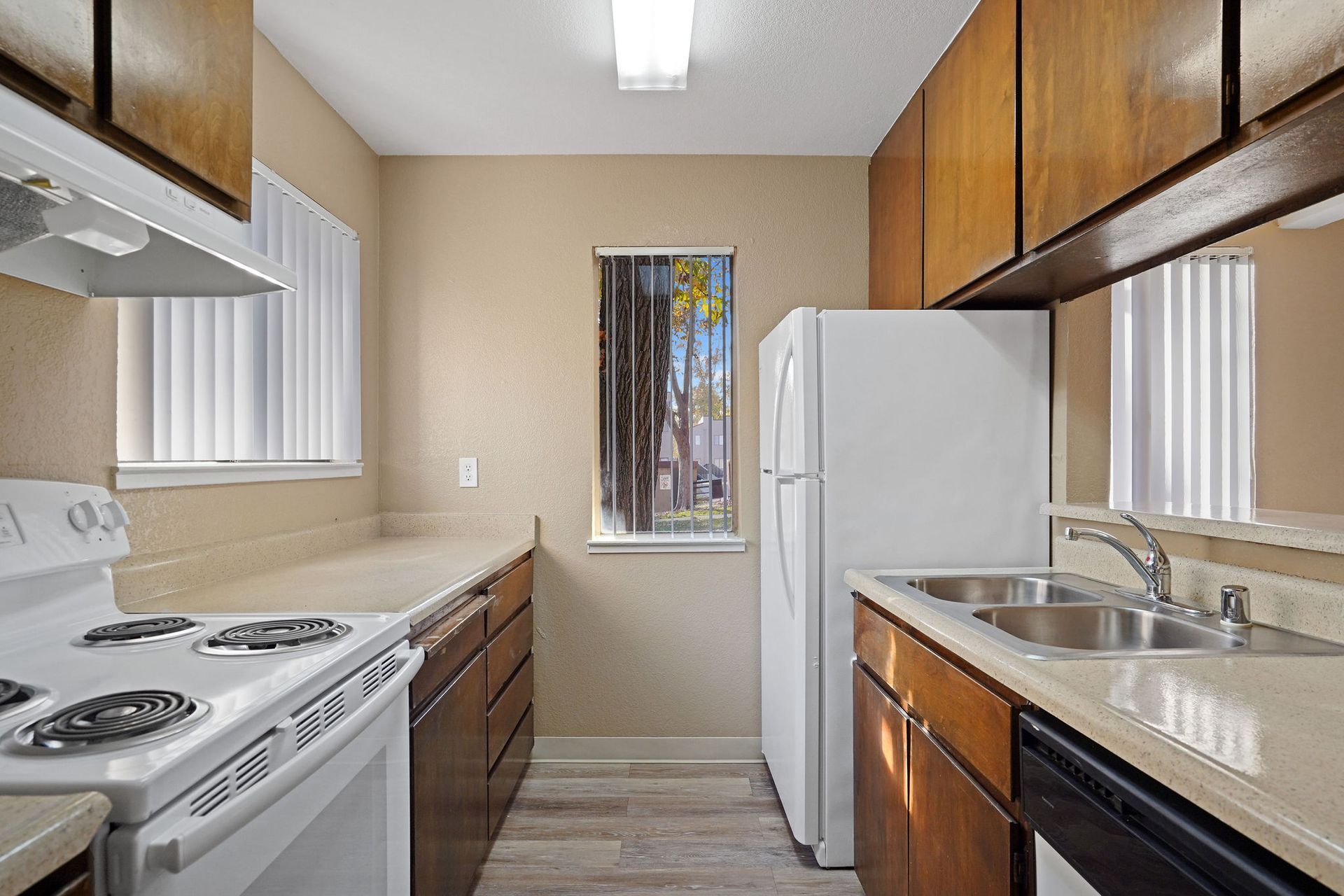 Narrow galley kitchen with white appliances, wood cabinets, and a sink by a bright window.