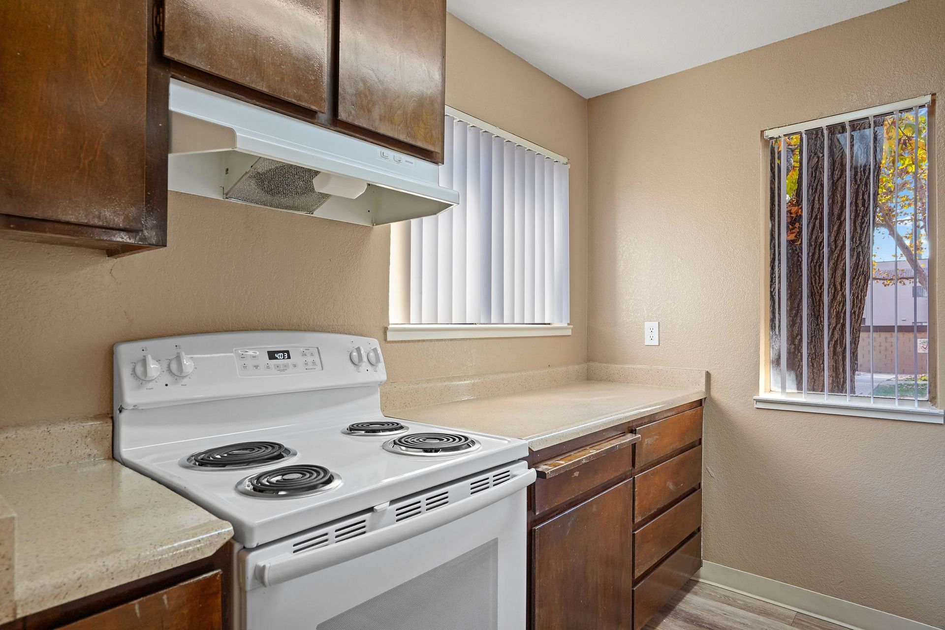 Small kitchen with white stove, beige counters, wooden cabinets, and two windows.