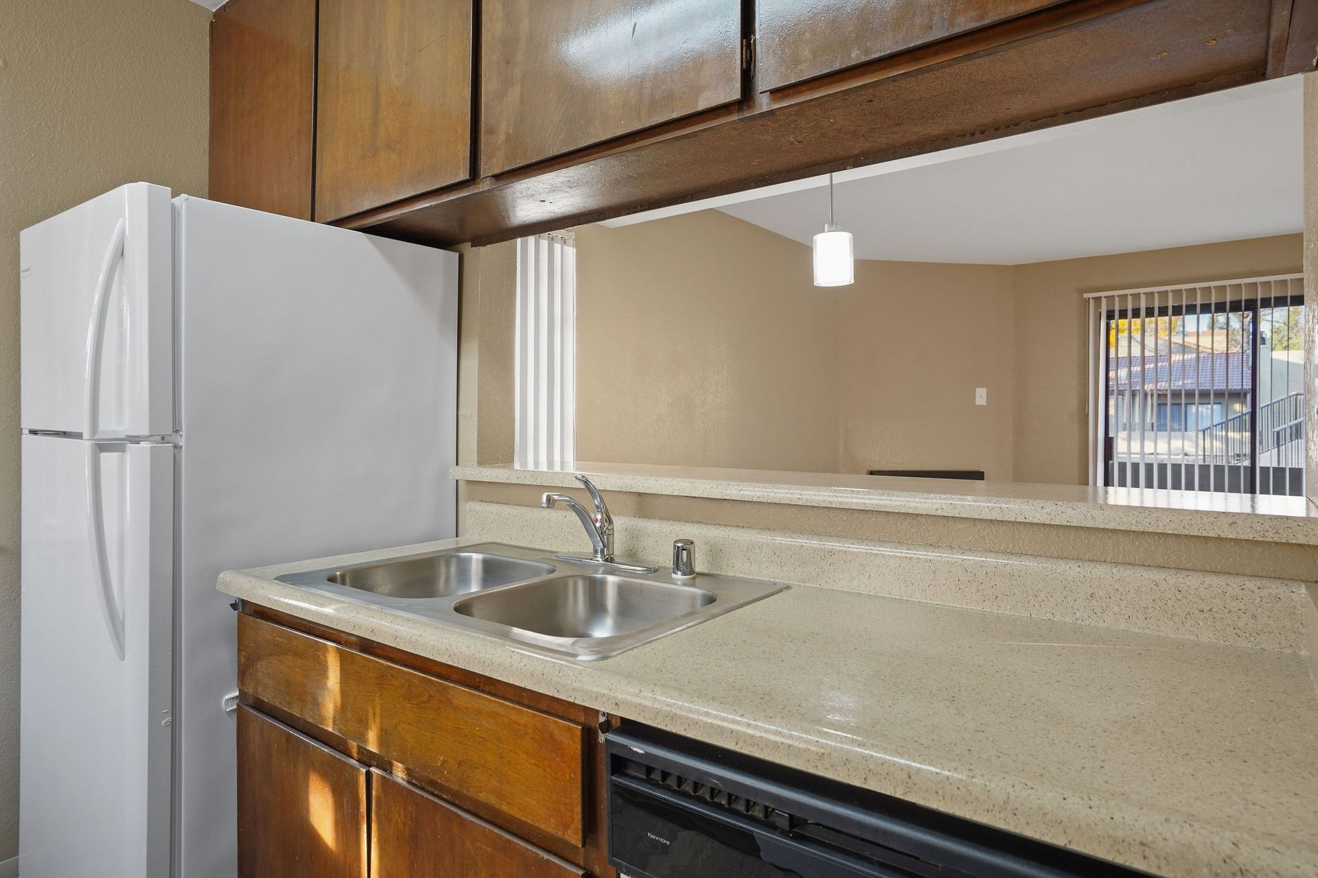 Kitchen with white refrigerator, sink, beige countertop, and wooden cabinets, viewed through a pass-through opening