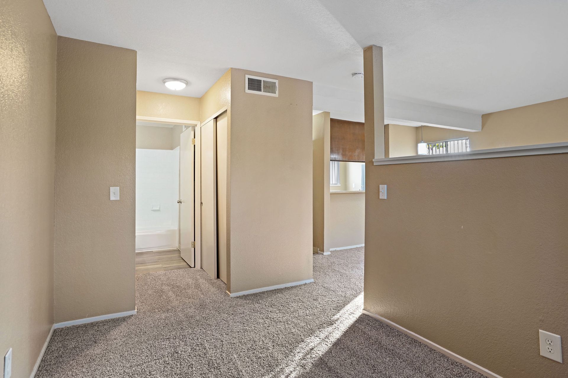 Empty beige apartment hallway and living area with carpeted floors and a partial view of the kitchen.
