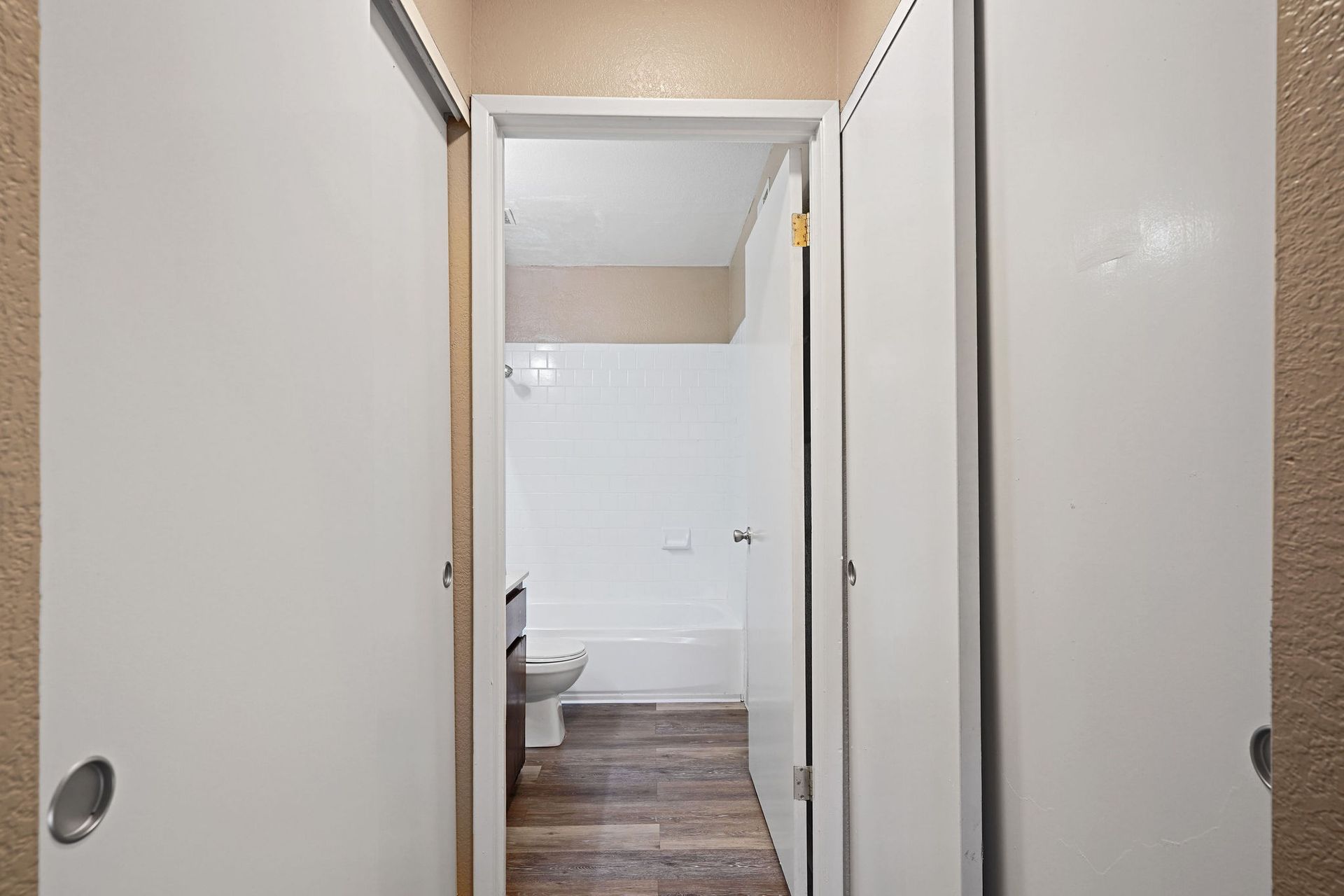 Narrow hallway with open door leading to a small bathroom with white walls and wood floor.