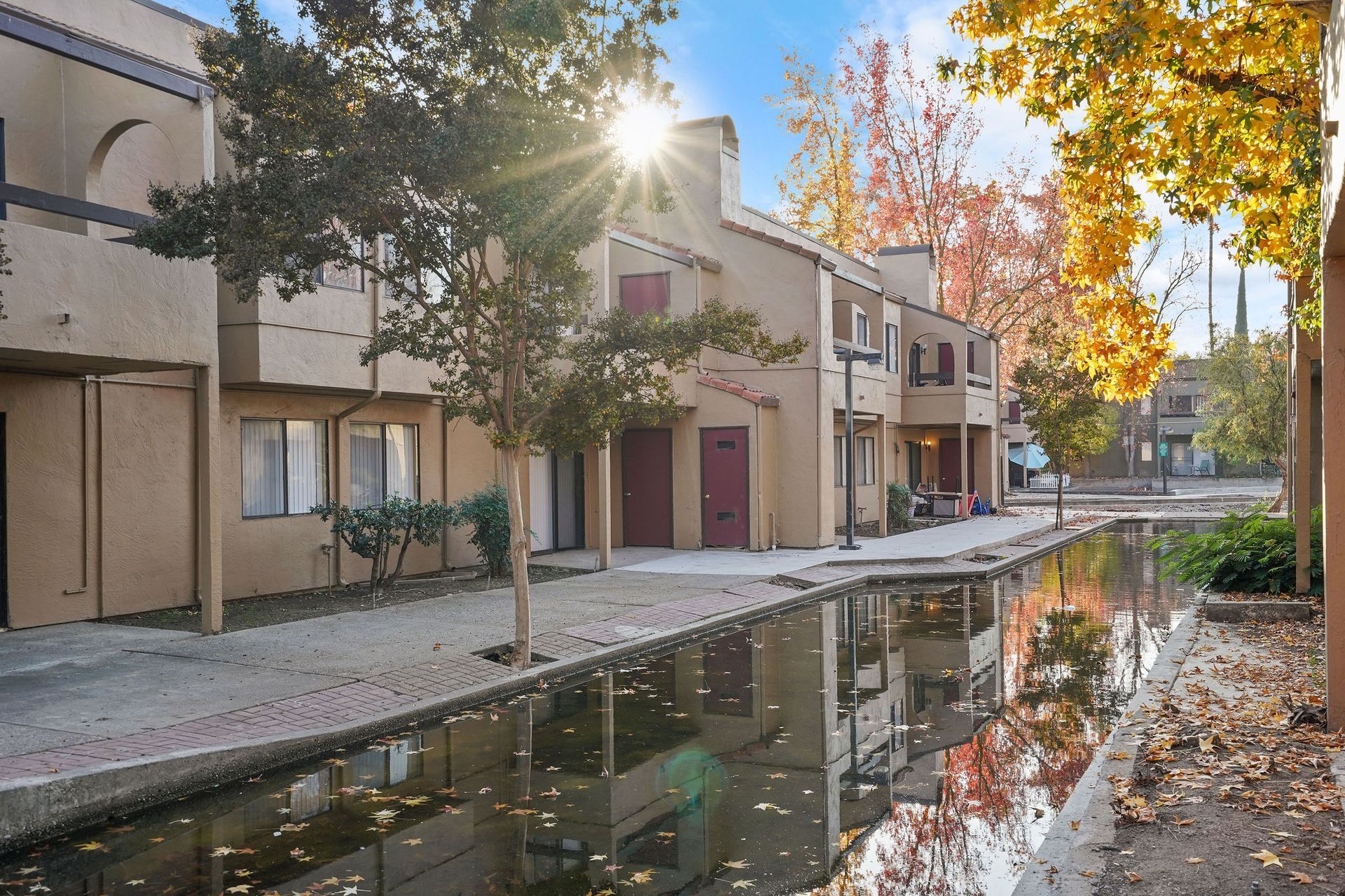 Sunlit courtyard with a narrow water canal beside tan townhouses and autumn trees