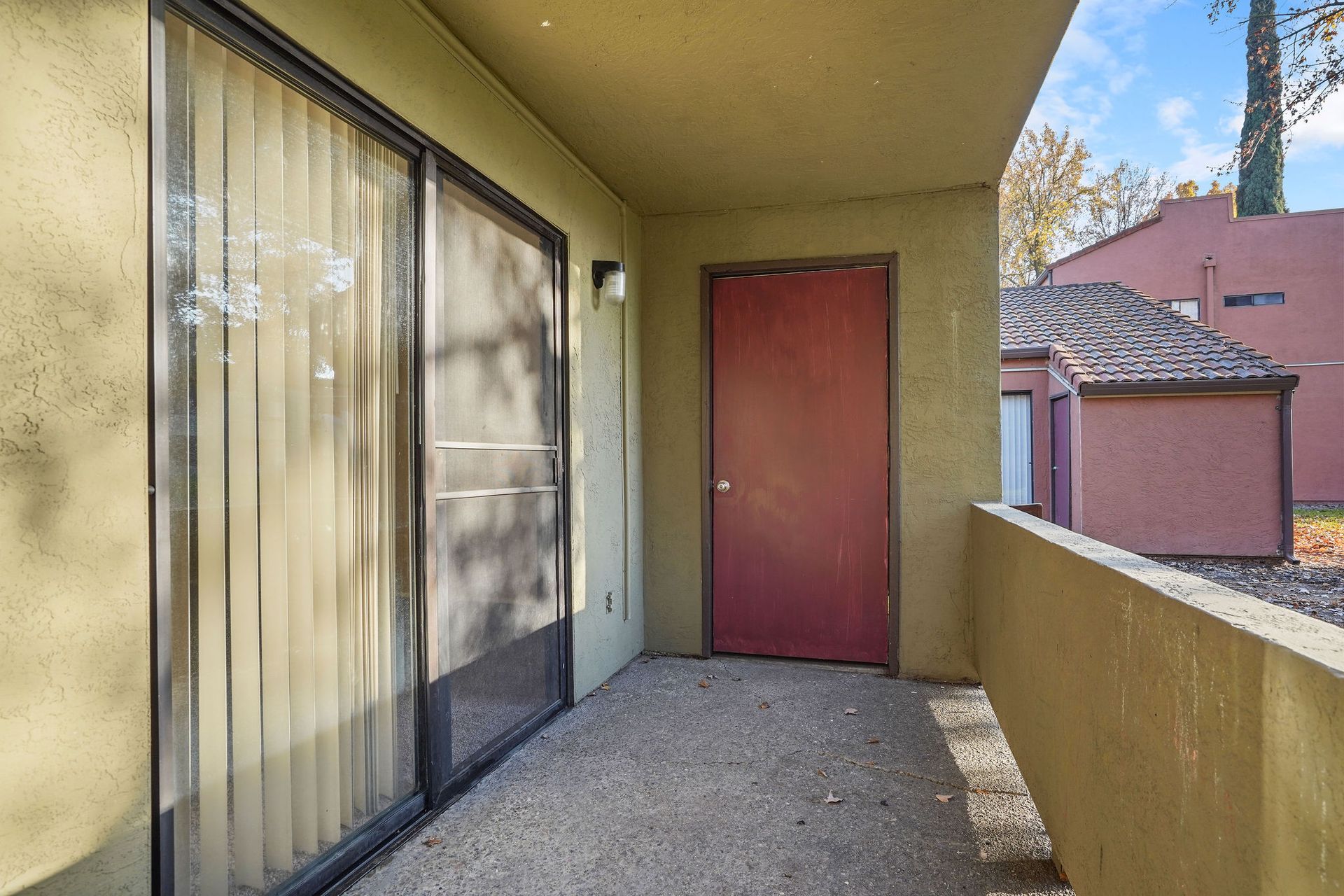 Covered apartment balcony with sliding glass door, red entry door, and beige stucco walls.