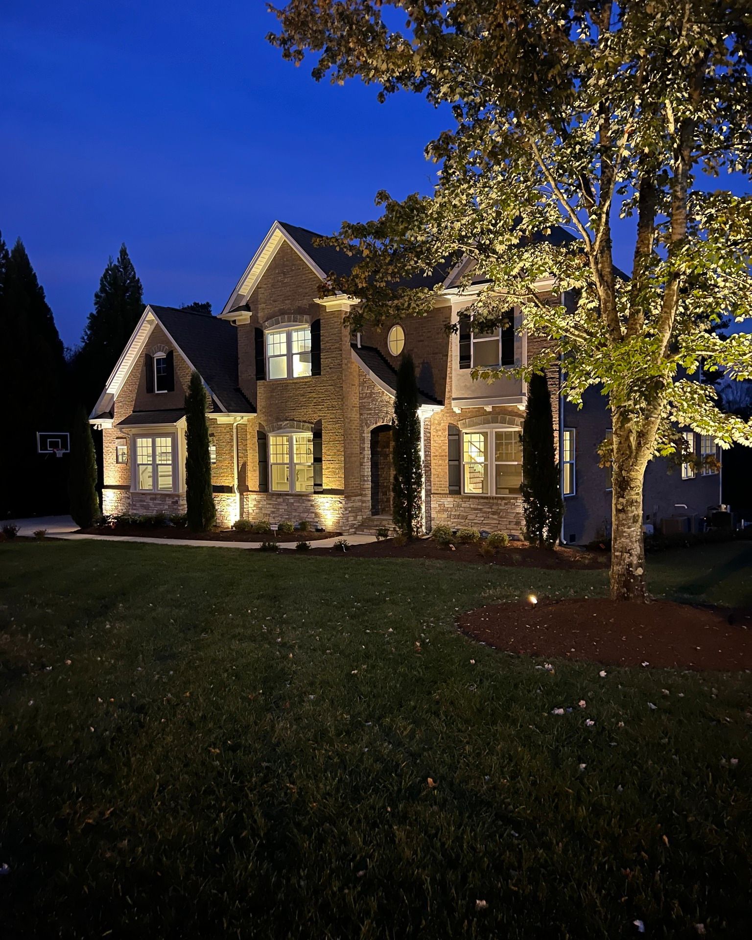 Two-story house with illuminated facade and tree at dusk; green lawn and dark blue sky.
