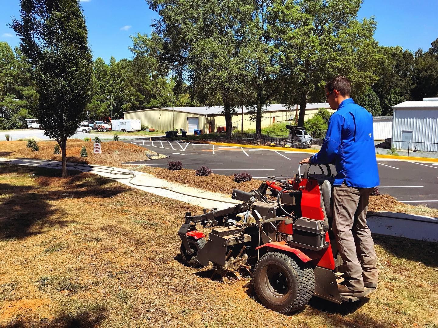 Man operating a small machine aerating a lawn in front of a parking area, trees in the background.