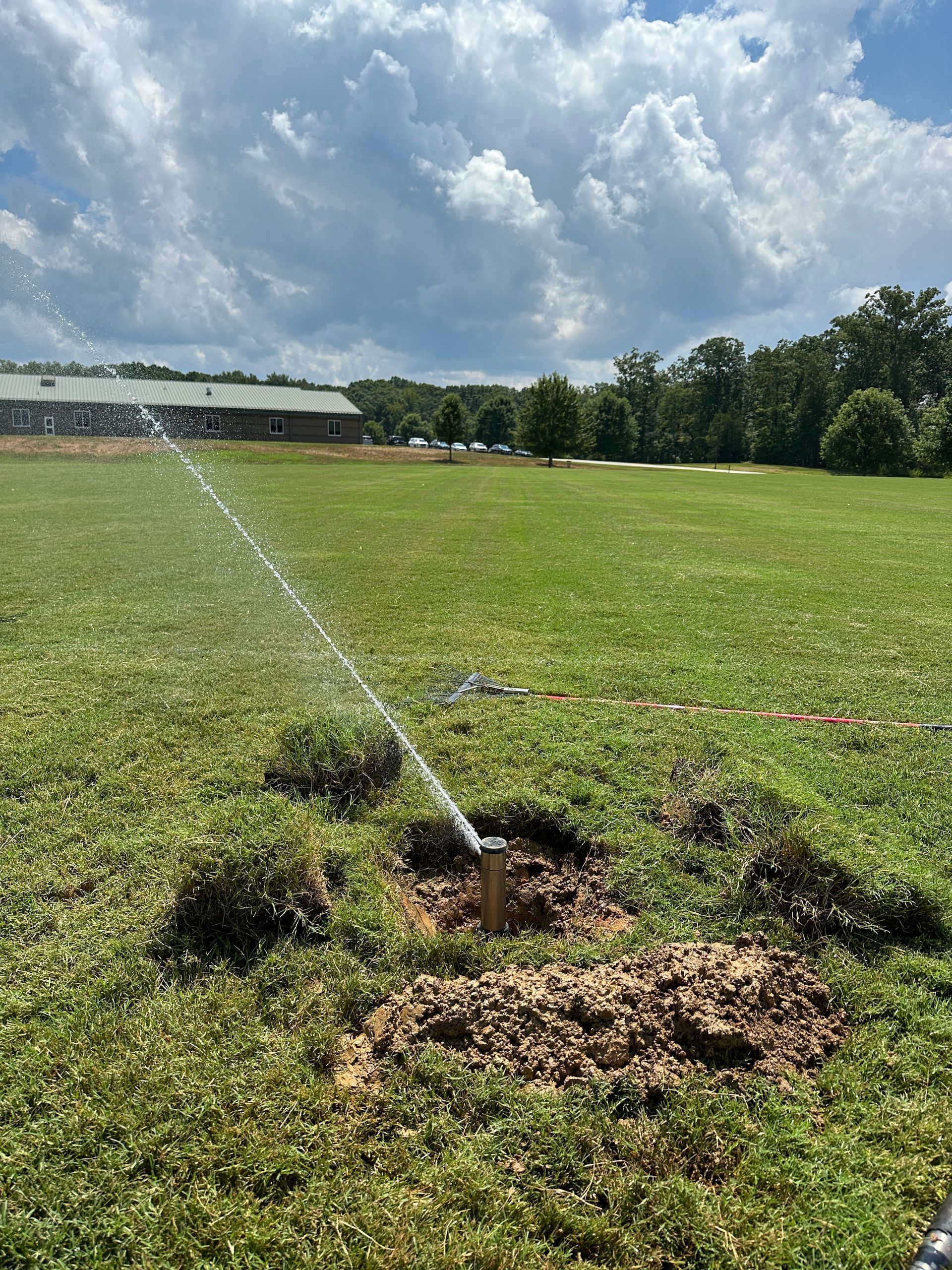 Sprinkler spraying water on a grassy field with a building in the background. Overcast sky.