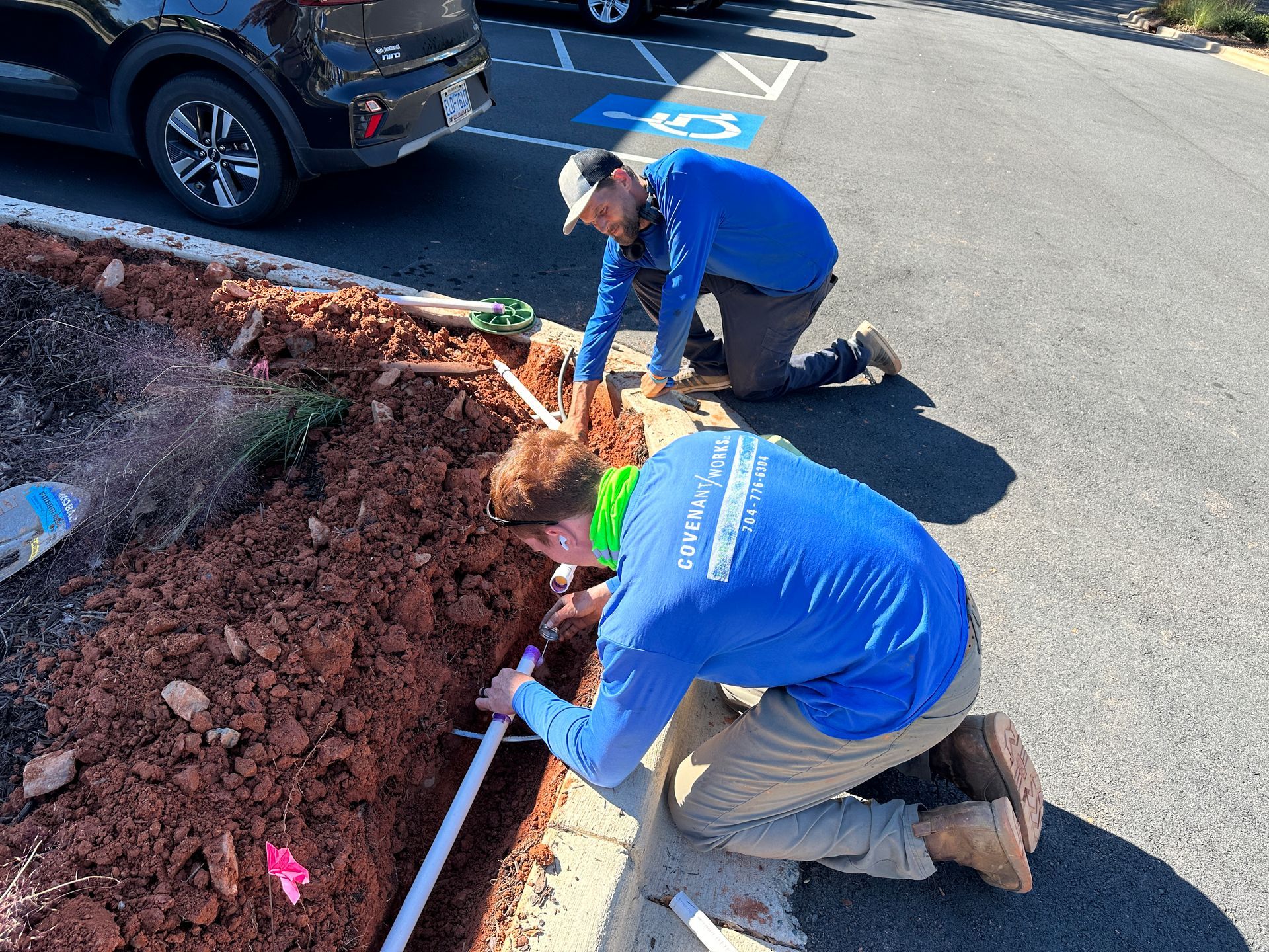 Two people in blue shirts install irrigation pipes near a curb by a parking lot.