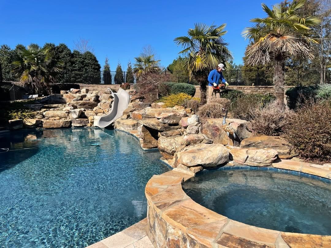 Swimming pool with rock waterfall and slide. Man stands near edge, palm trees and blue sky.