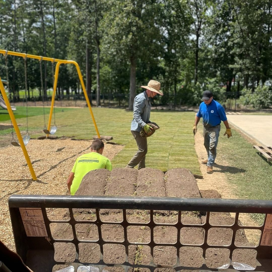 Workers laying sod near a playground. Two men carry grass rolls, one unloads sod. Yellow swing set is nearby.
