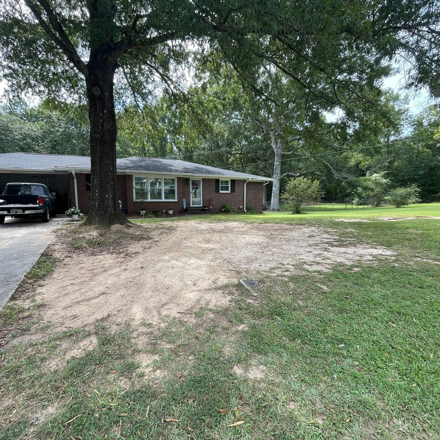 House with a brick facade, driveway, and dirt patch in the front yard; tree provides shade.