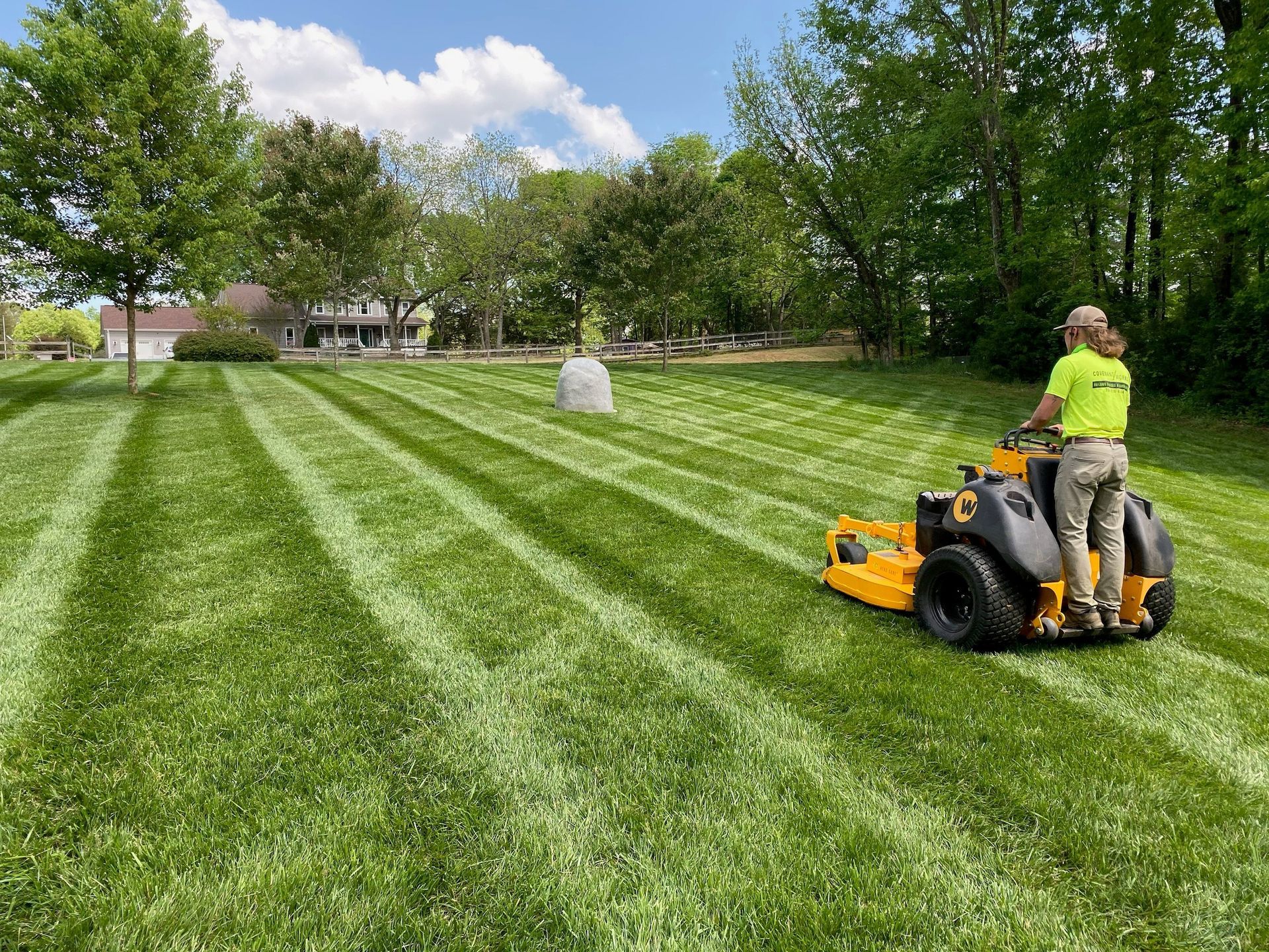 A person mowing a lawn with a riding mower, leaving striped patterns on the grass.