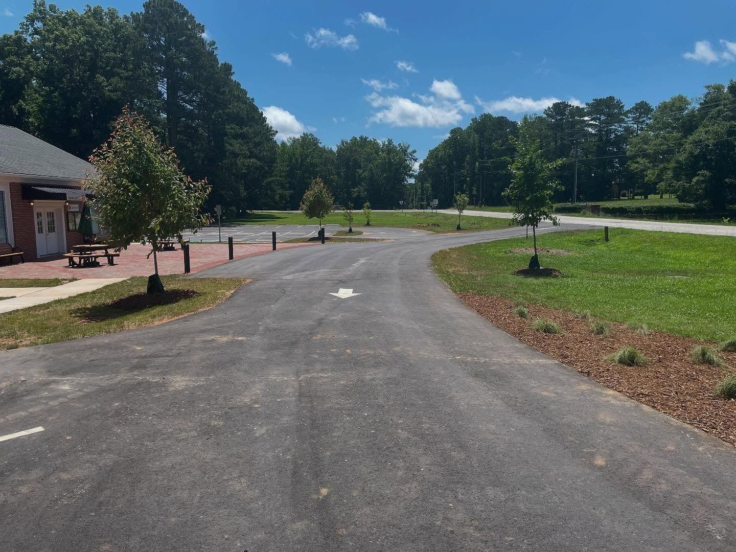 Paved road with arrow leads toward trees and grass. Building on left. Blue sky with clouds.