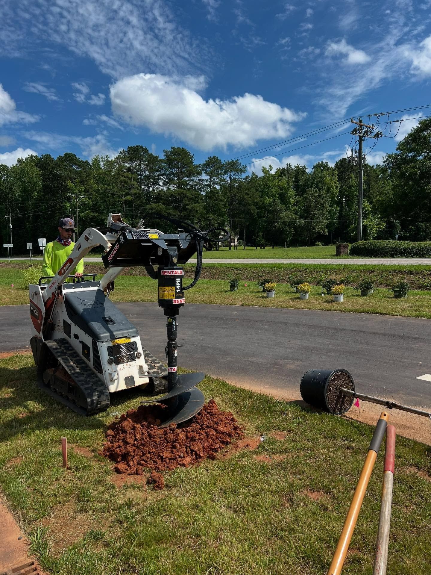 A person operating a Bobcat auger to dig a hole on a grassy area, with a potted plant nearby.