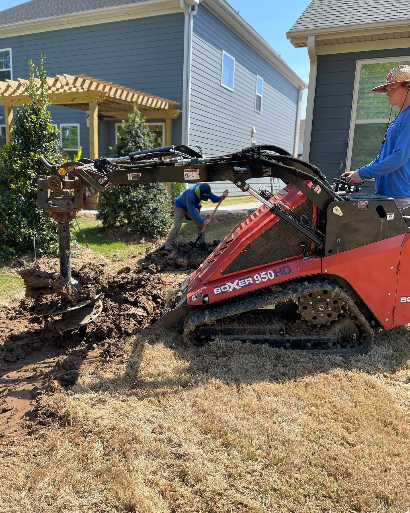 Mini skid steer with auger digging in yard, another person shoveling; two houses and a pergola in the background.