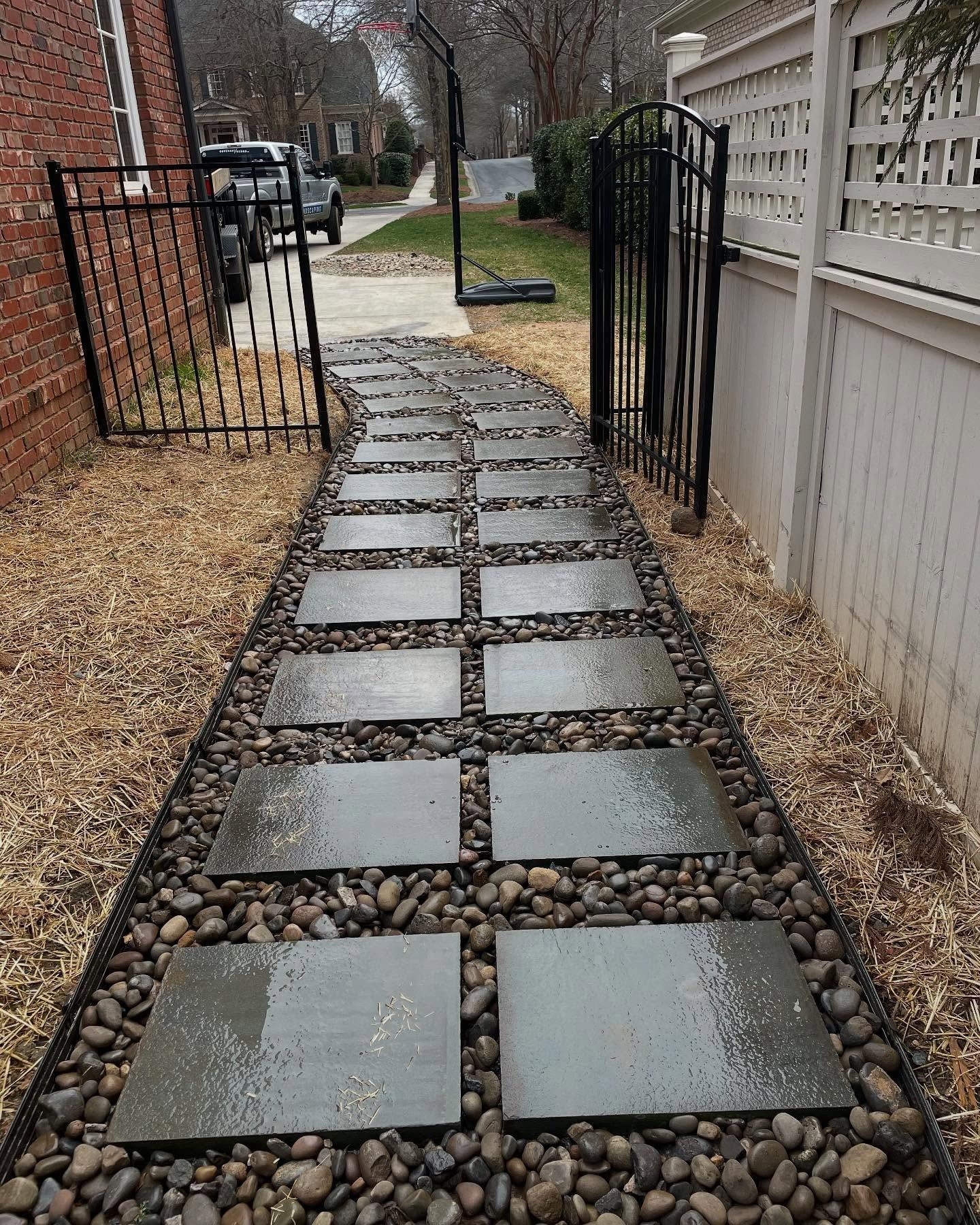 Stone path with square pavers through a rock bed, next to black fence and white fence.