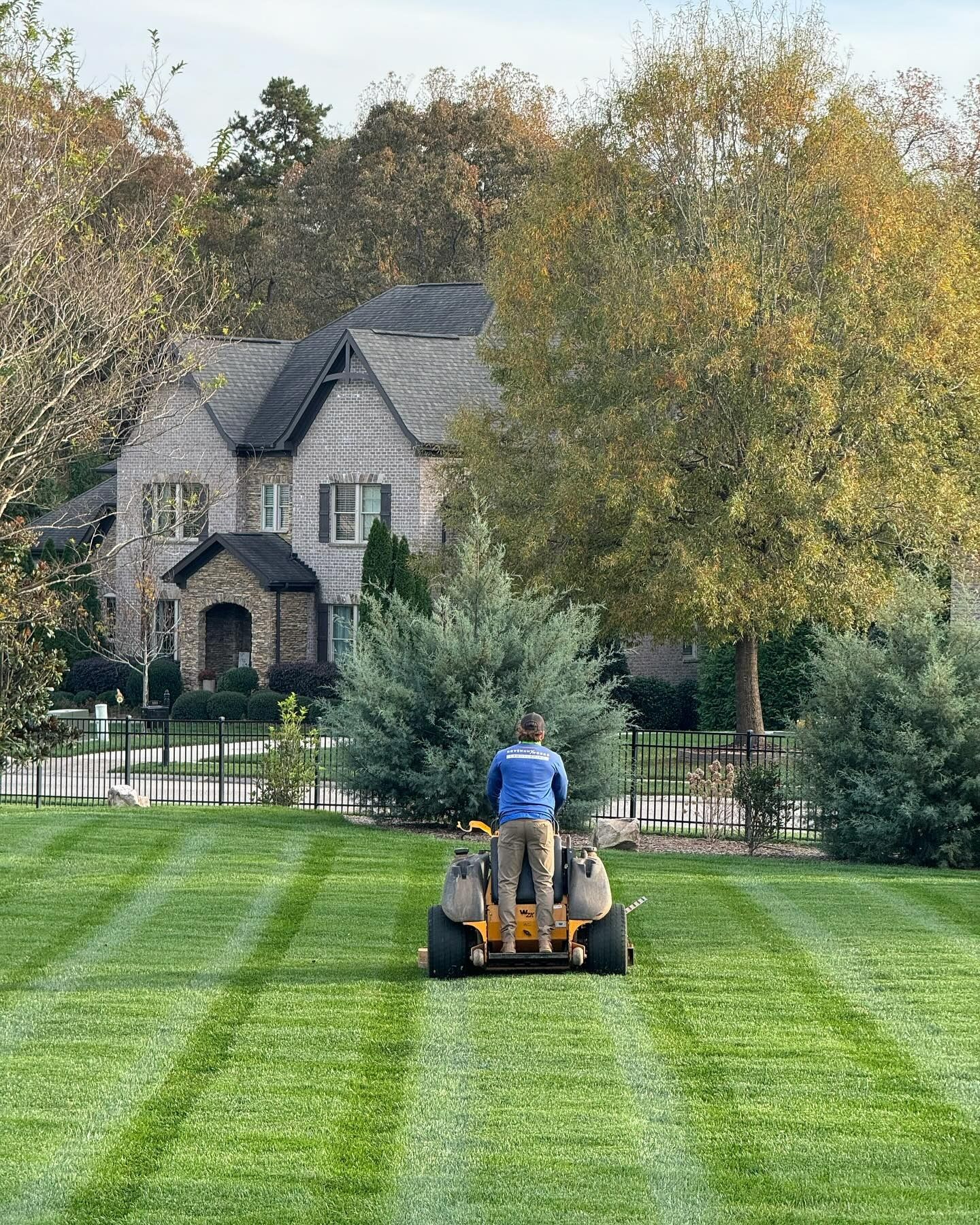 Man mowing a striped green lawn in front of a large house with a stone entryway.