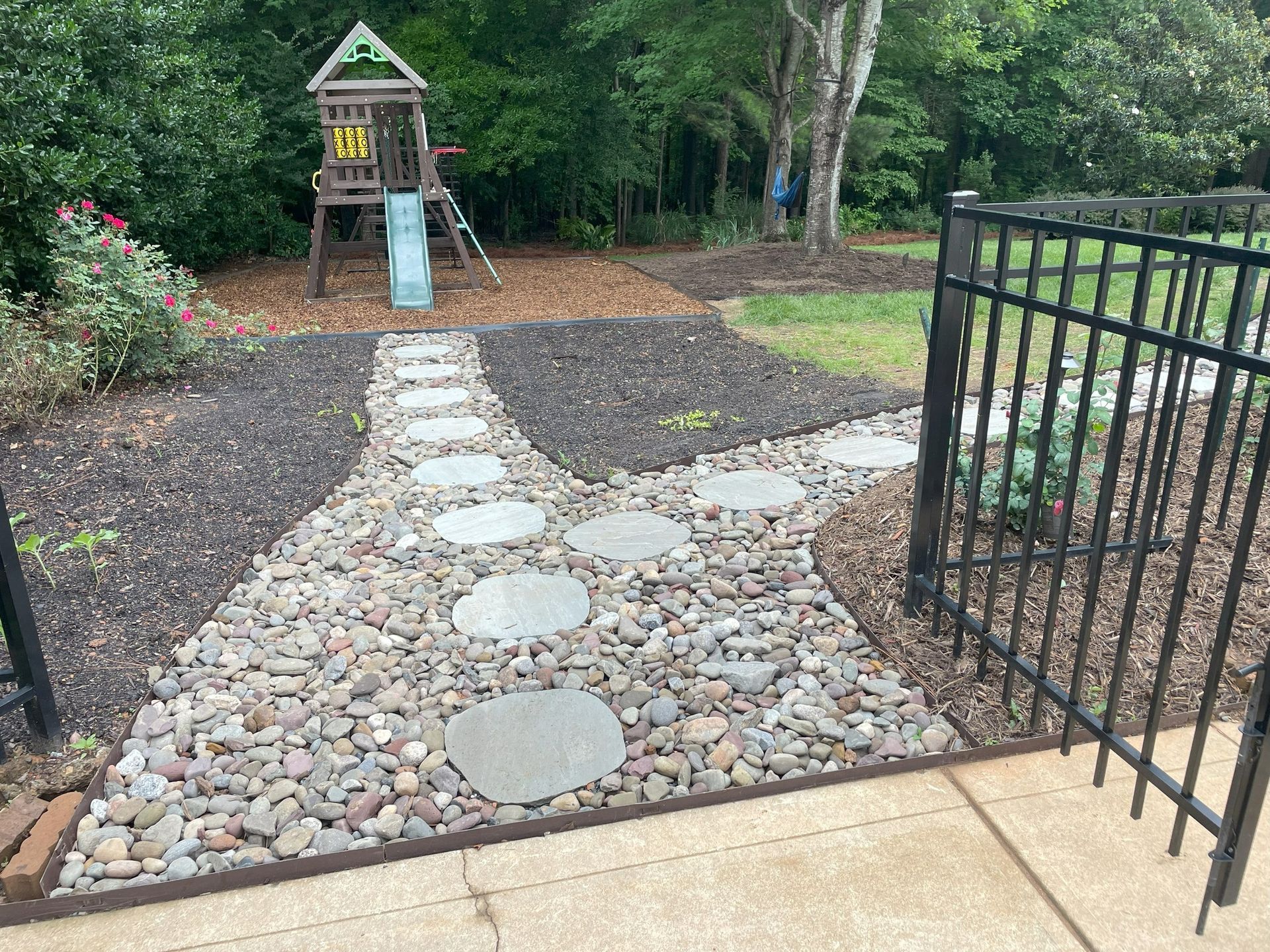 Backyard with a stone path leading to a wooden play structure and gate. Stone and mulch ground cover.