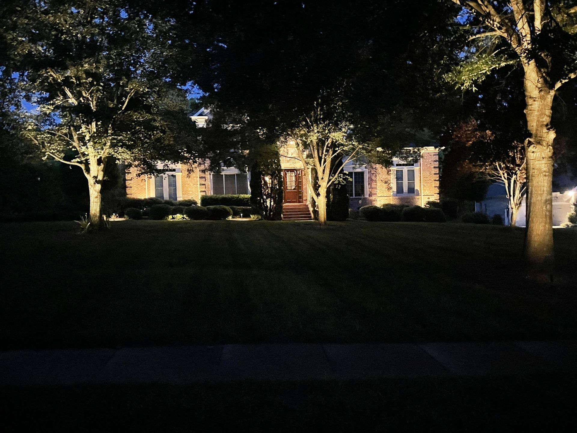 House at night with landscaping lights illuminating the facade, lawn, and surrounding trees.