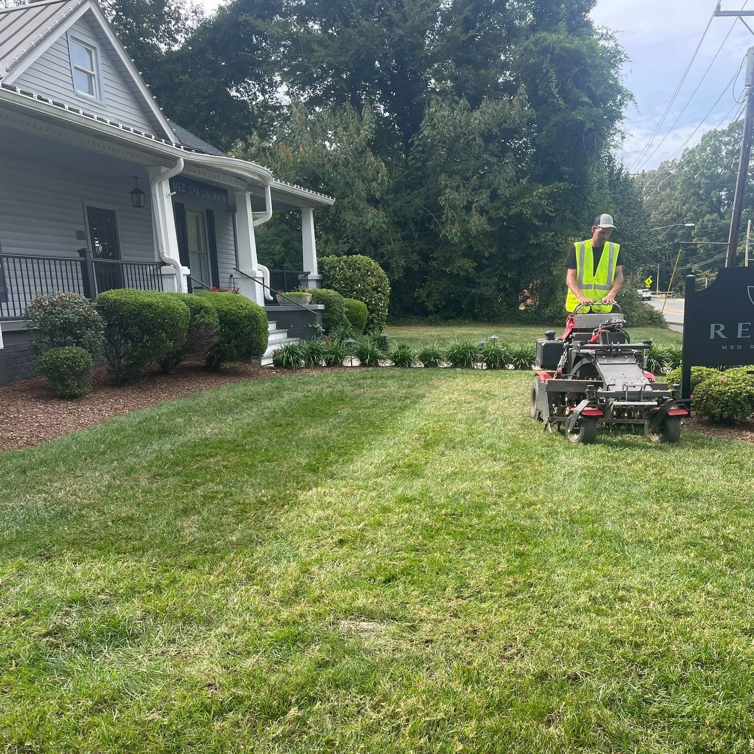 Man in safety vest mowing a lawn with a riding mower next to a house.