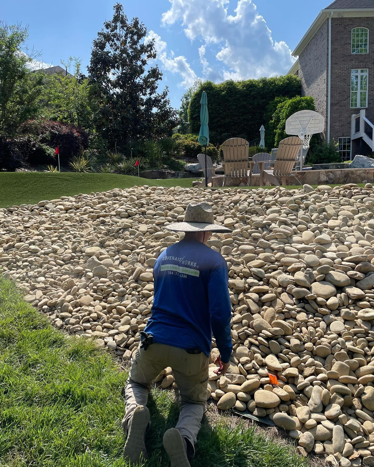 Person kneeling on the grass, inspecting a large rock-covered drainage area. Sunny day, residential setting.