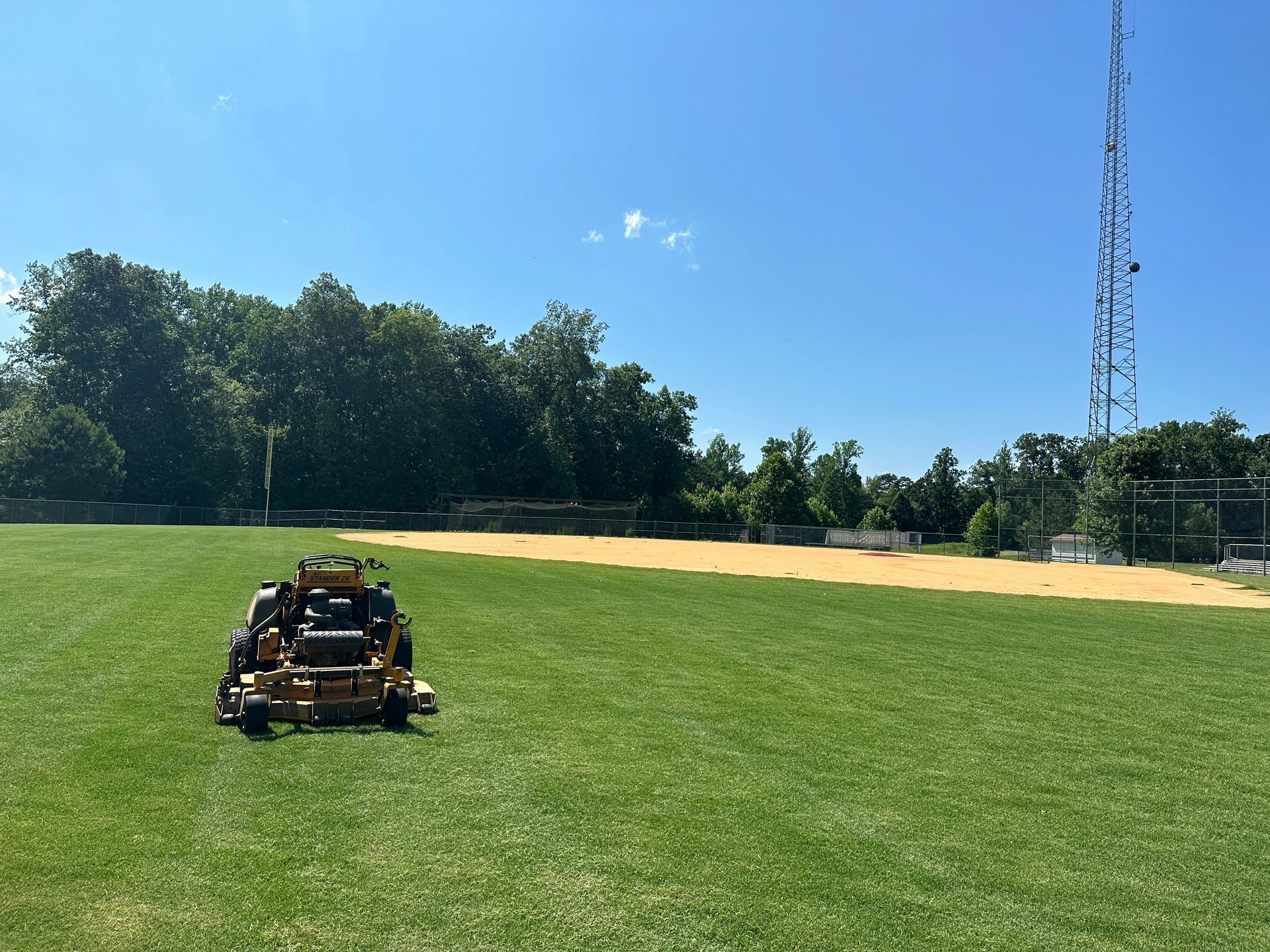 A riding lawn mower cuts grass on a sunny field near a baseball diamond and treeline.