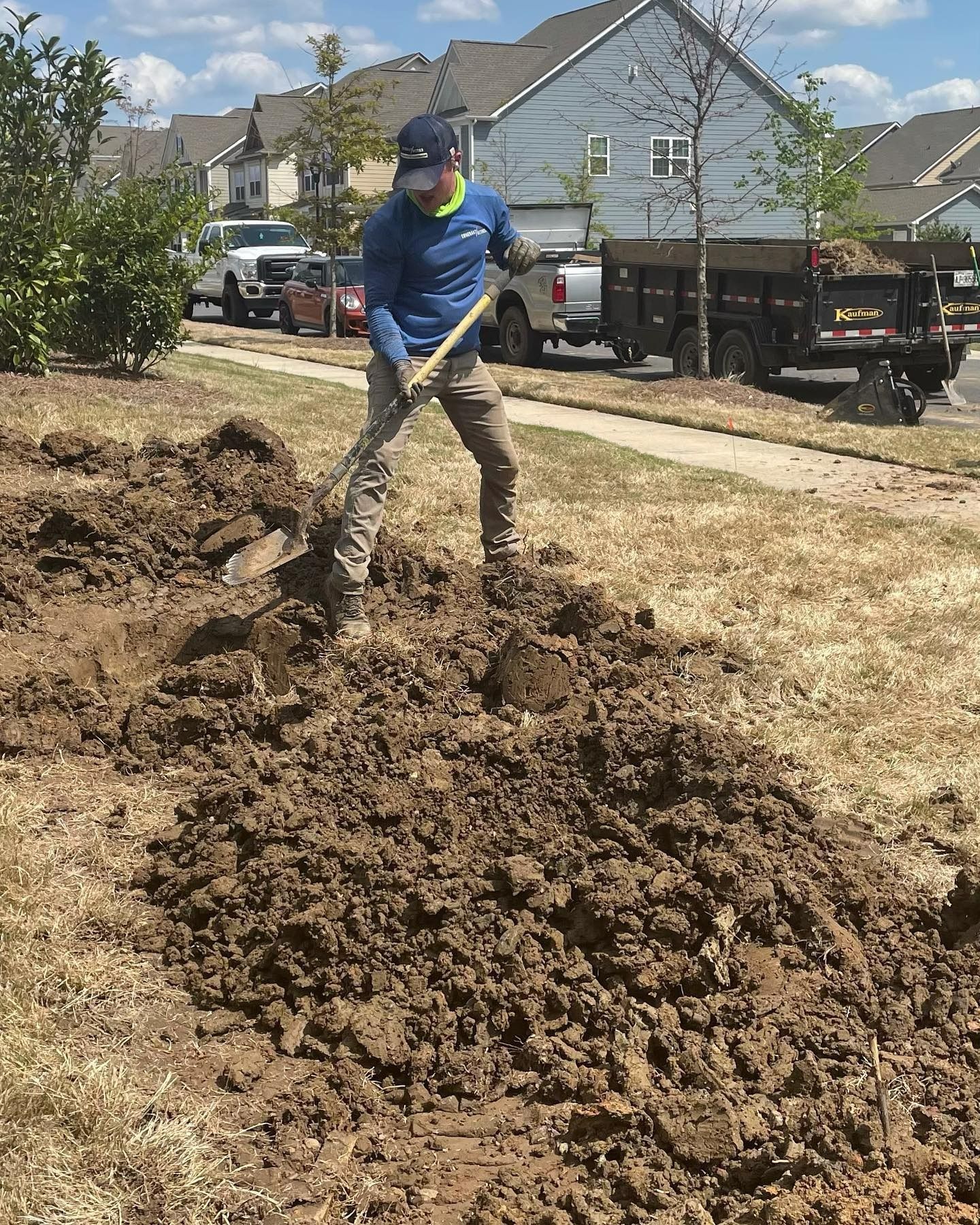 Man digging in dirt on a grassy verge, with a shovel.  A truck and houses are in the background.