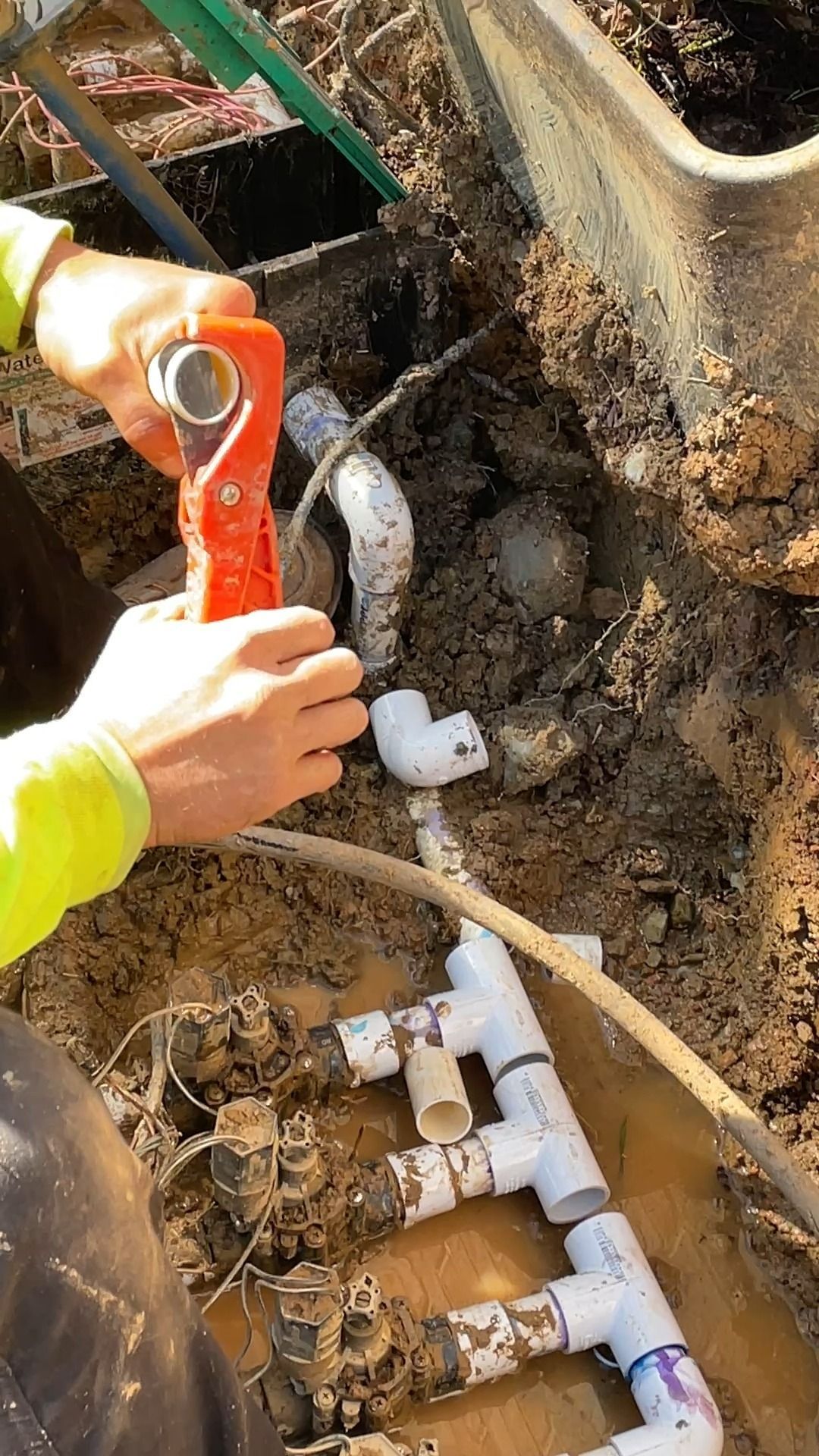 Plumber repairs buried PVC pipes, using a cutter in an excavated area outdoors, wearing a neon green shirt.