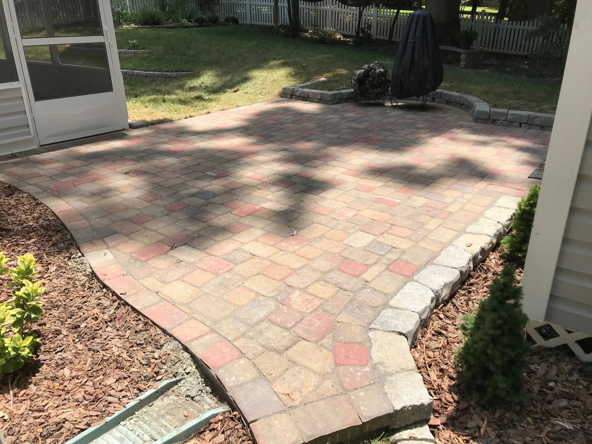 Brick patio with curved edges, surrounded by mulch and small plants, next to a house and screened porch.