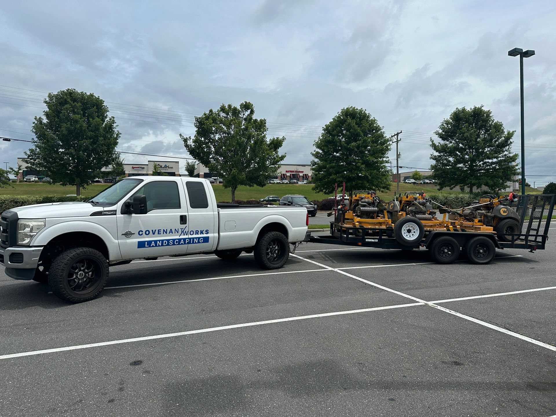 White pickup truck towing a trailer with landscaping equipment in a parking lot.