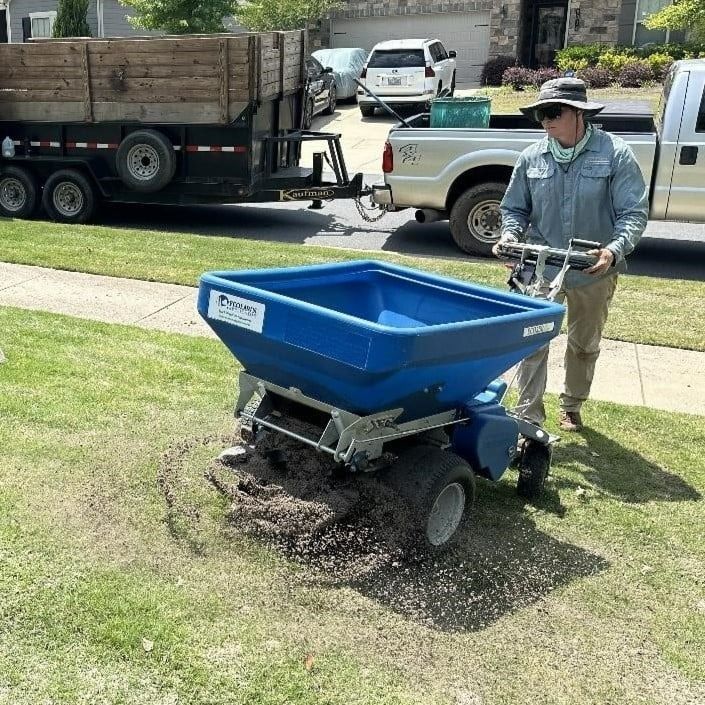 Man using a blue spreader on a lawn, trailer and truck in the background.