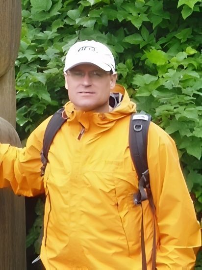 Man in yellow jacket and white cap, backpack, standing outdoors near green foliage.
