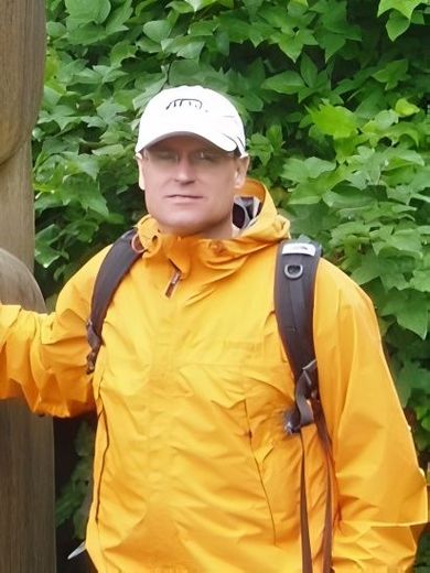 Man in yellow jacket and white cap, backpack, standing outdoors near green foliage.