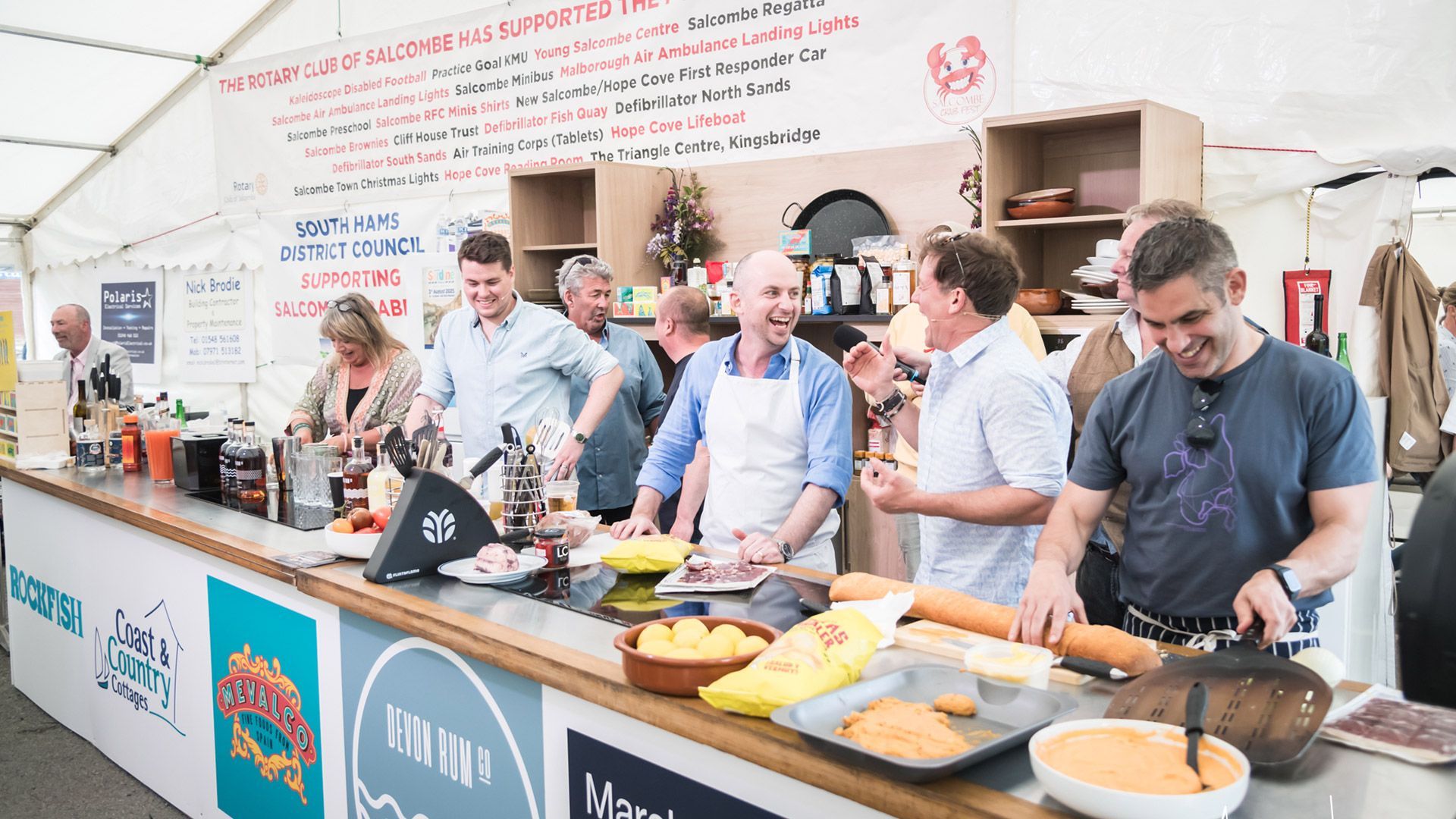 People at a food event cooking behind a long counter, smiling.